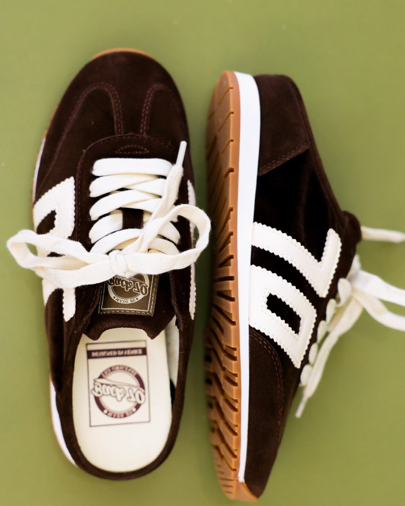 A pair of vintage-style low-top sneakers in dark brown suede with white stripes, white laces, and gum soles, displayed on a solid olive green background.