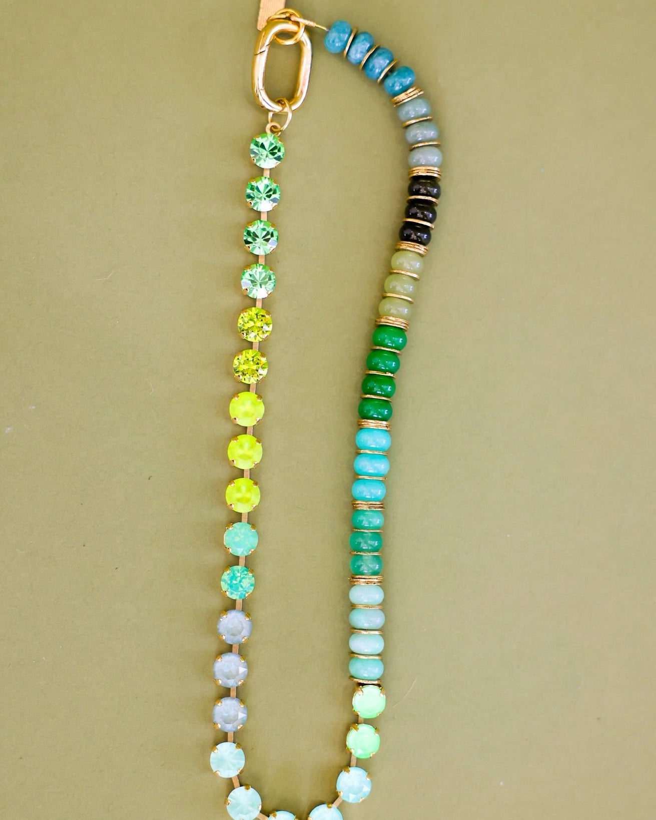 A beaded bracelet with a gradient of colorful round beads transitioning from pastel blue and purple to green, yellow, and turquoise, finished with a gold clasp on a solid beige background.
