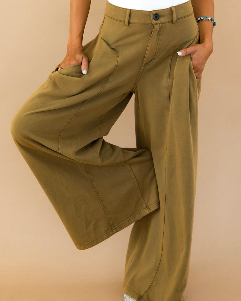 A woman posing in a white short-sleeve top and olive green wide-leg pants against a solid beige studio background.