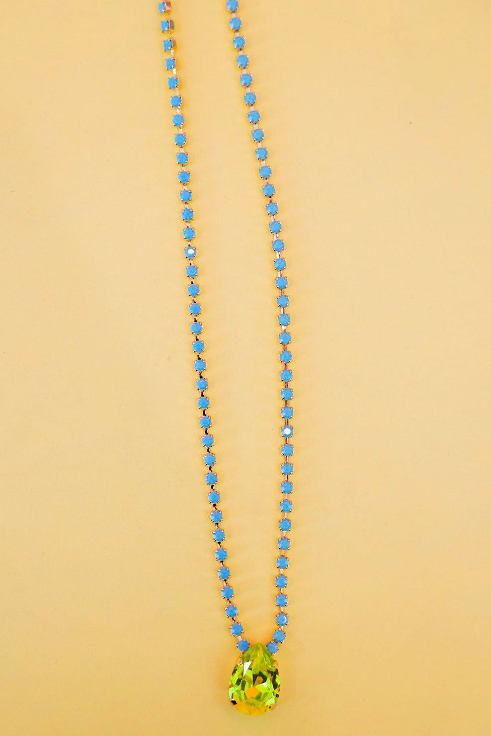A beaded necklace featuring small light blue faceted beads and a large, faceted lime green teardrop pendant, displayed against a solid pale peach background.