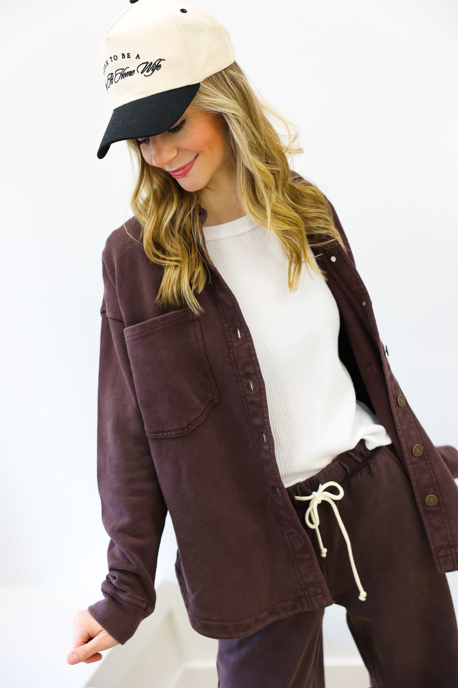 Woman wearing a brown jacket, white shirt, and purple pants with a beige cap.