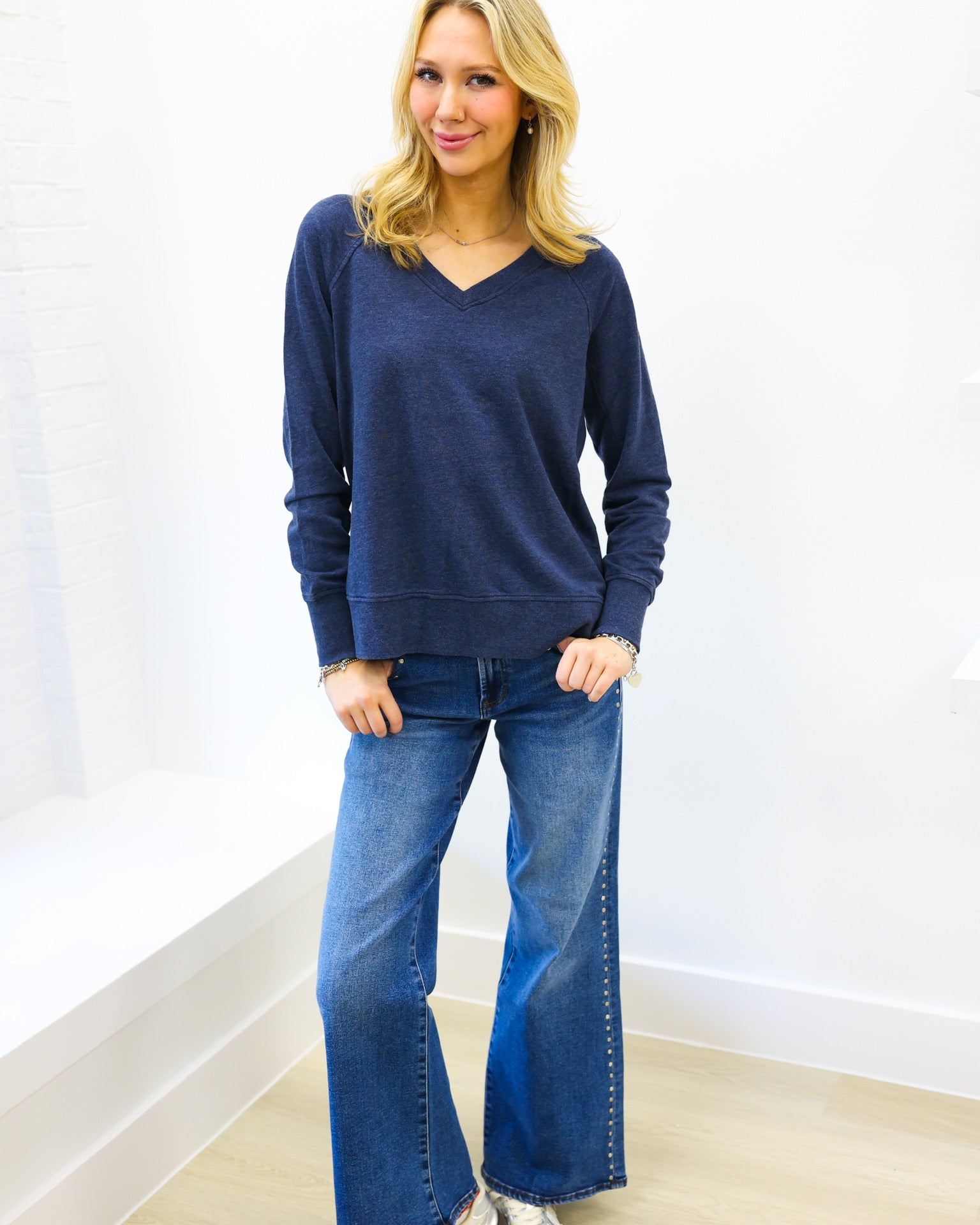 Woman wearing a blue sweater and jeans standing against a white wall.