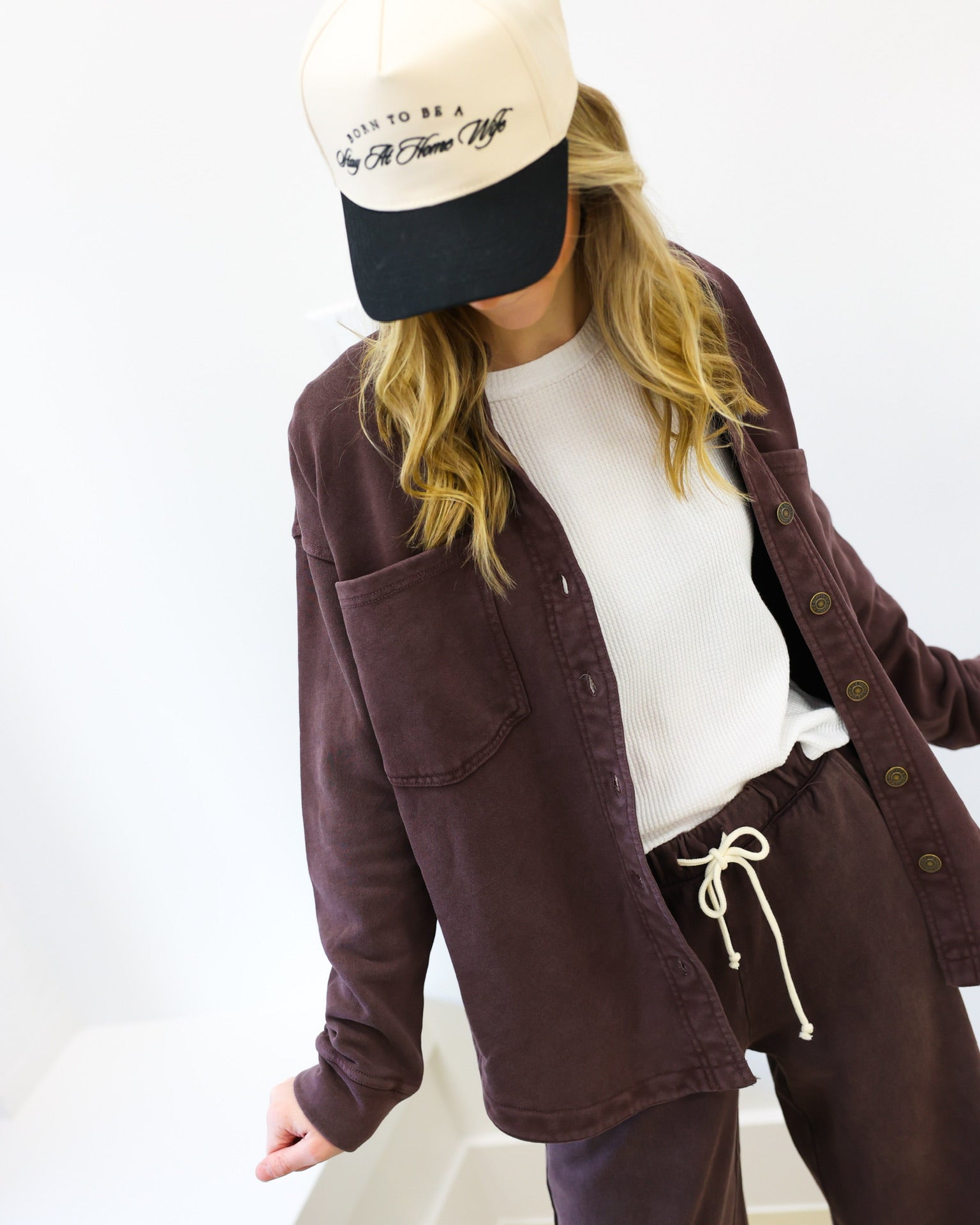 Person wearing a brown jacket, white shirt, and purple pants with a cap on a white background