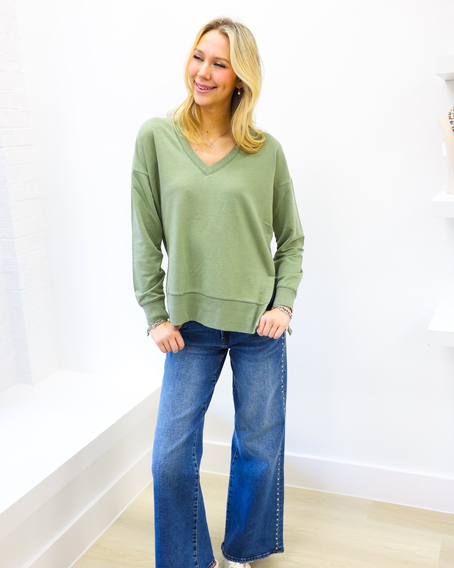 Woman wearing a green sweater and blue jeans standing against a white wall.