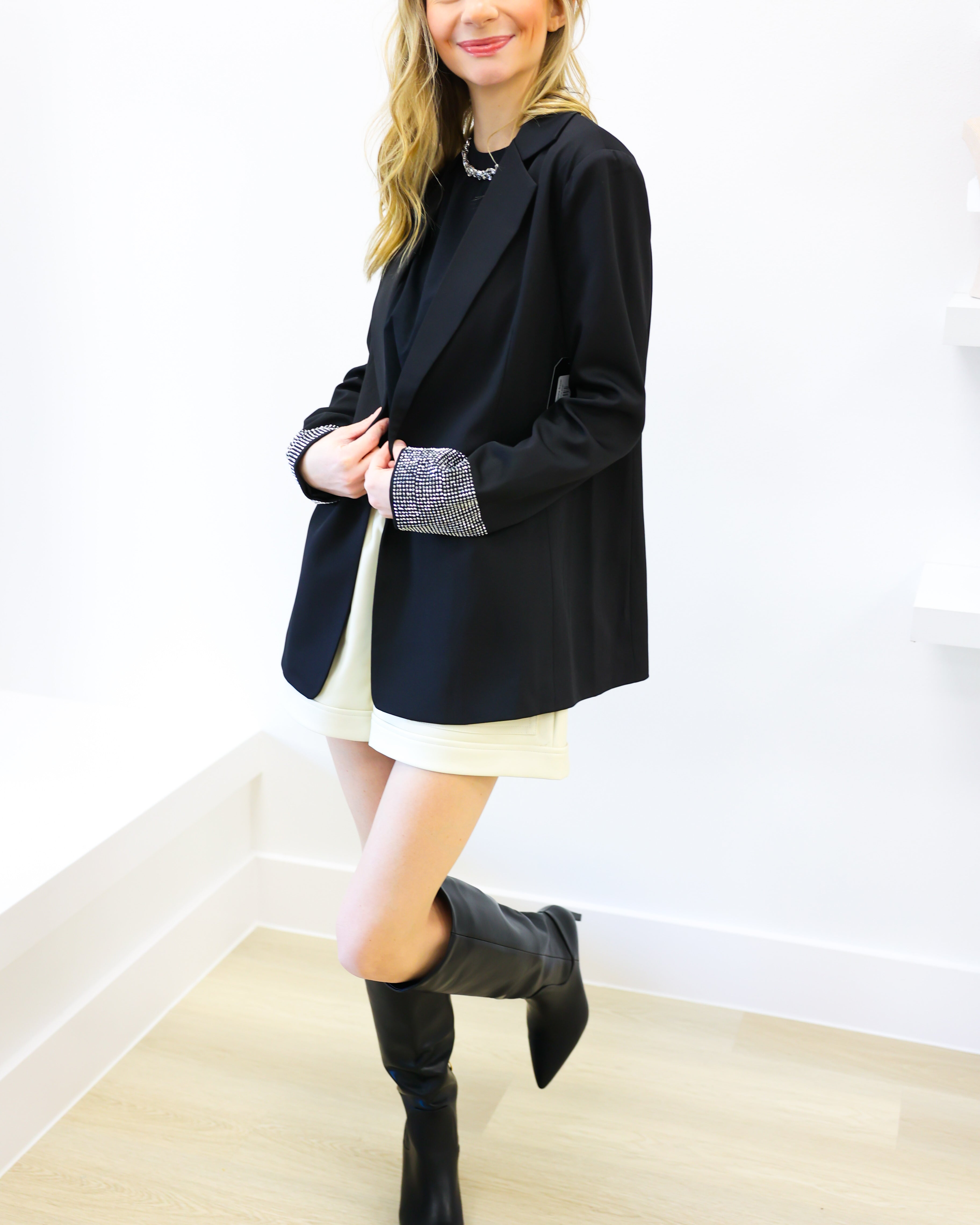 Woman wearing a black blazer, white dress, and black boots against a white wall.