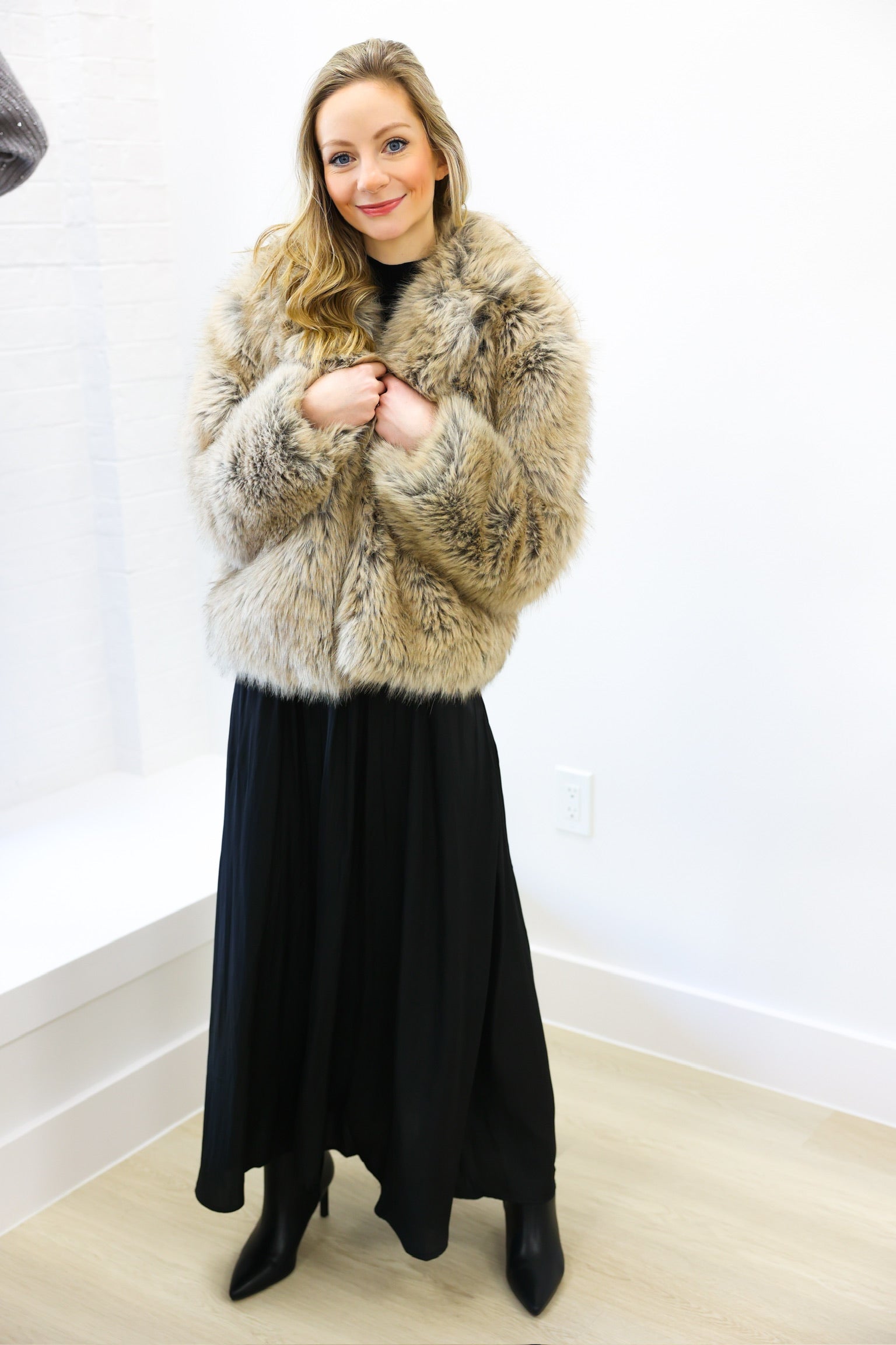 Woman wearing a fur coat and black dress indoors against a white wall.