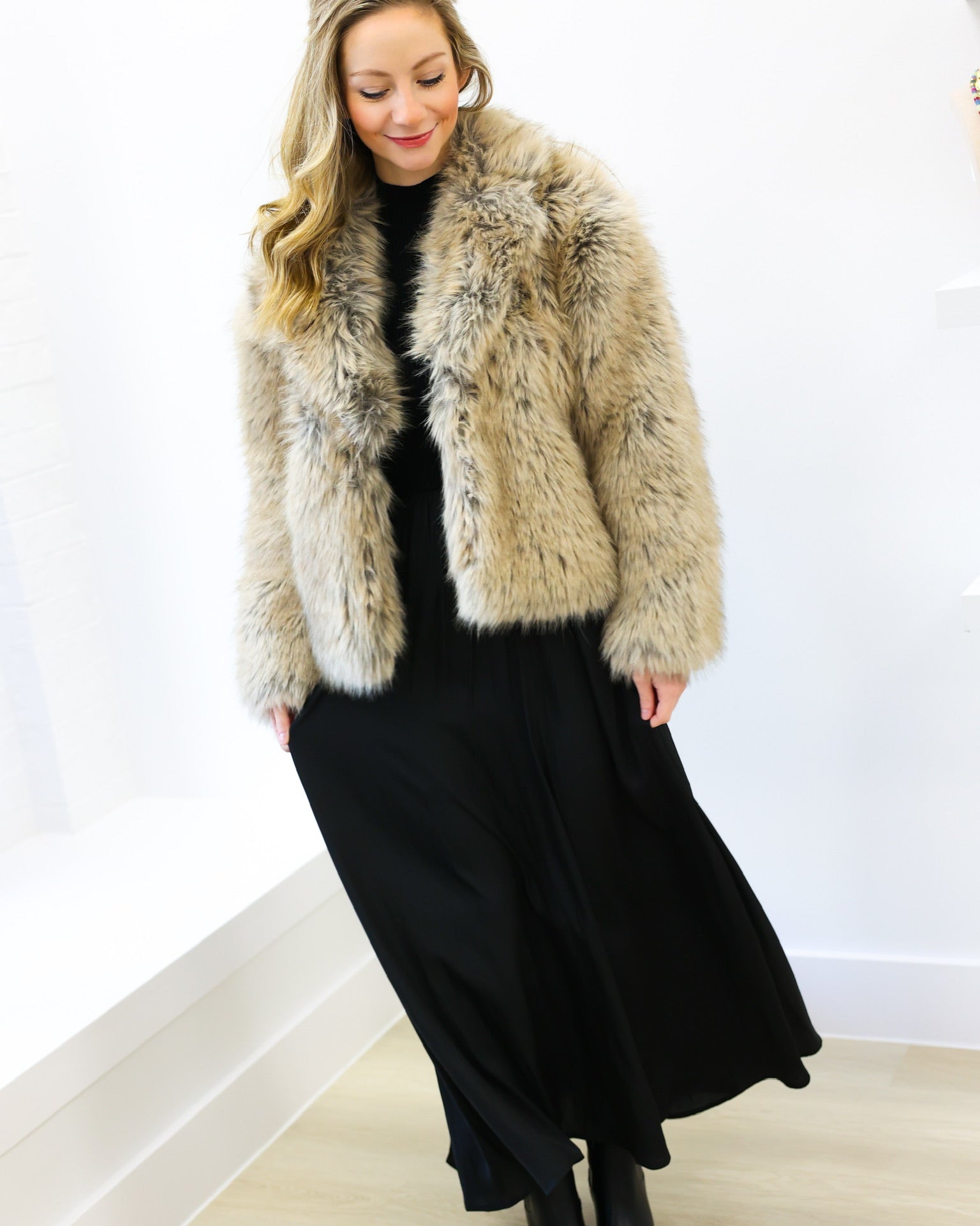 Woman wearing a fur coat over a black dress against a white wall.
