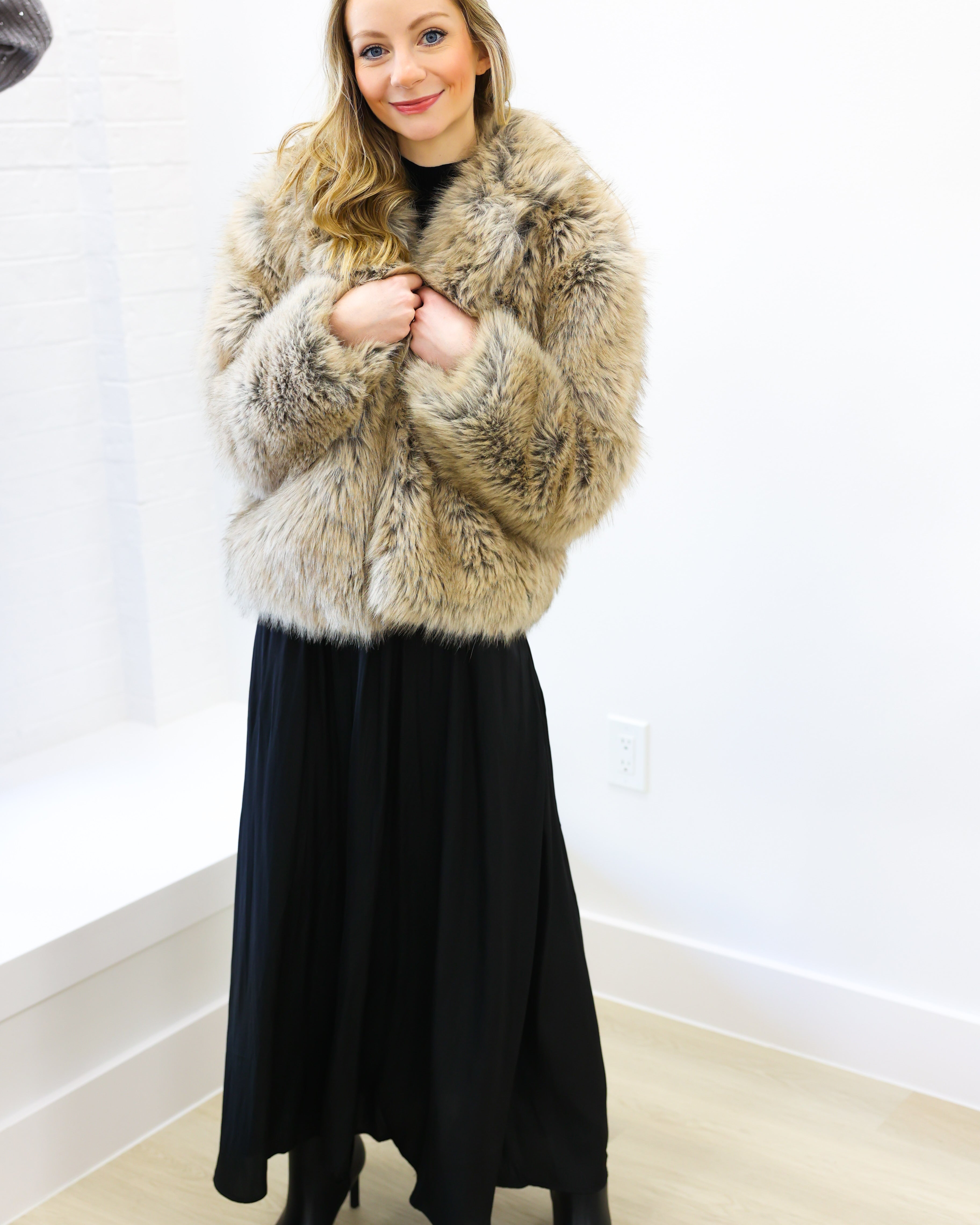 Woman wearing a fur coat and black dress indoors against a white wall.