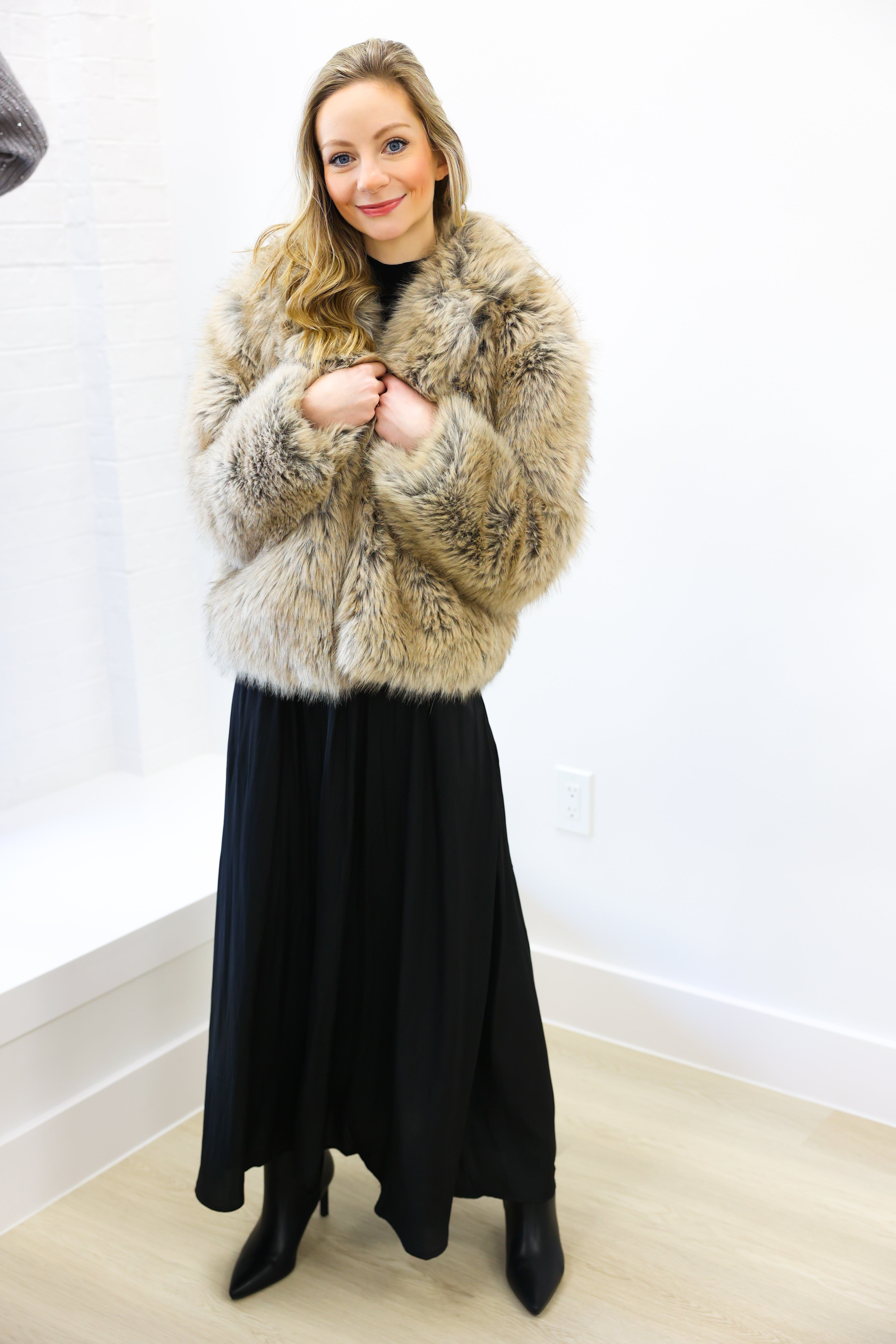 Woman wearing a fur coat and black dress indoors against a white wall.