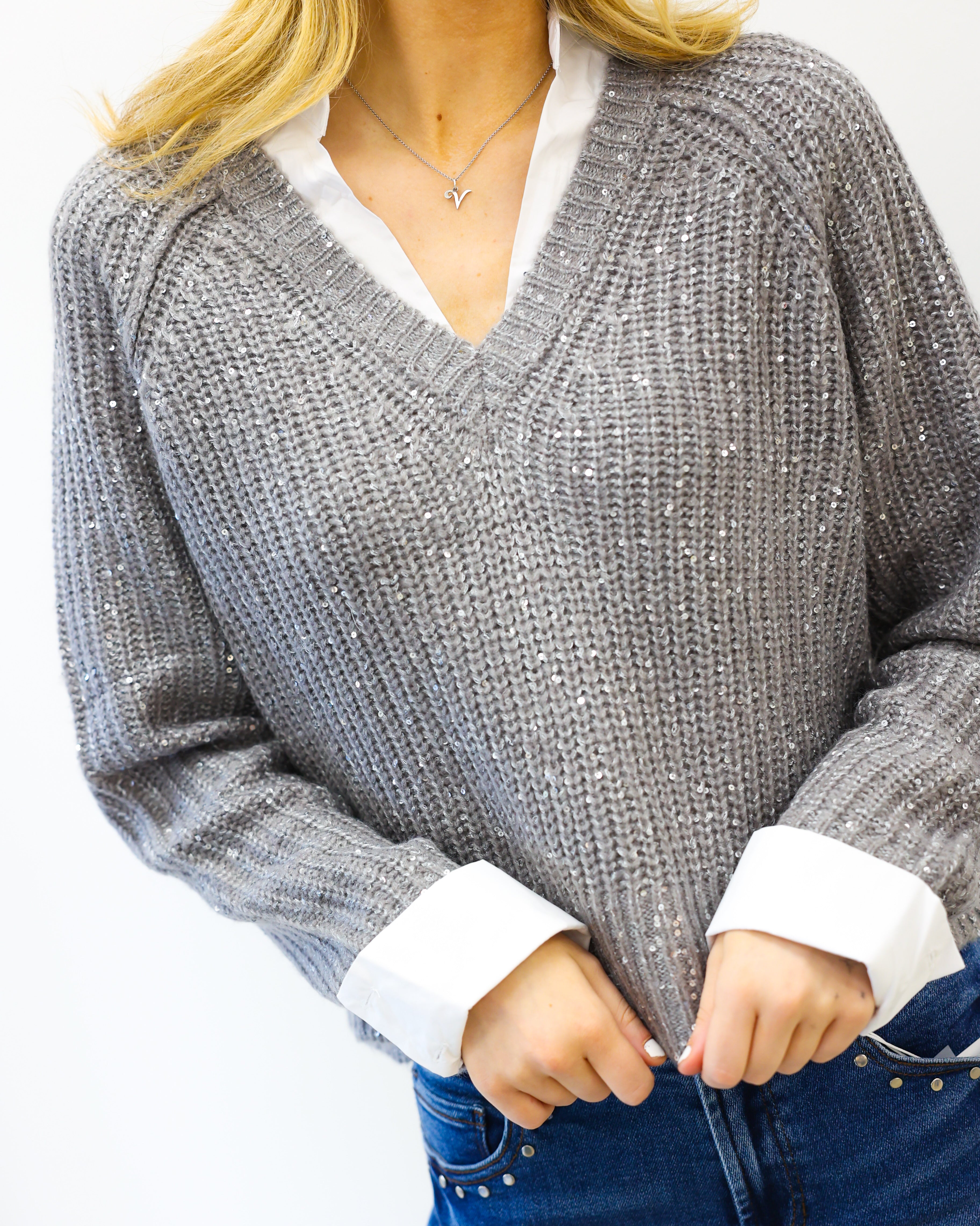 Person wearing a gray sweater with white cuffs and collar, blue jeans, and a necklace on a white background.