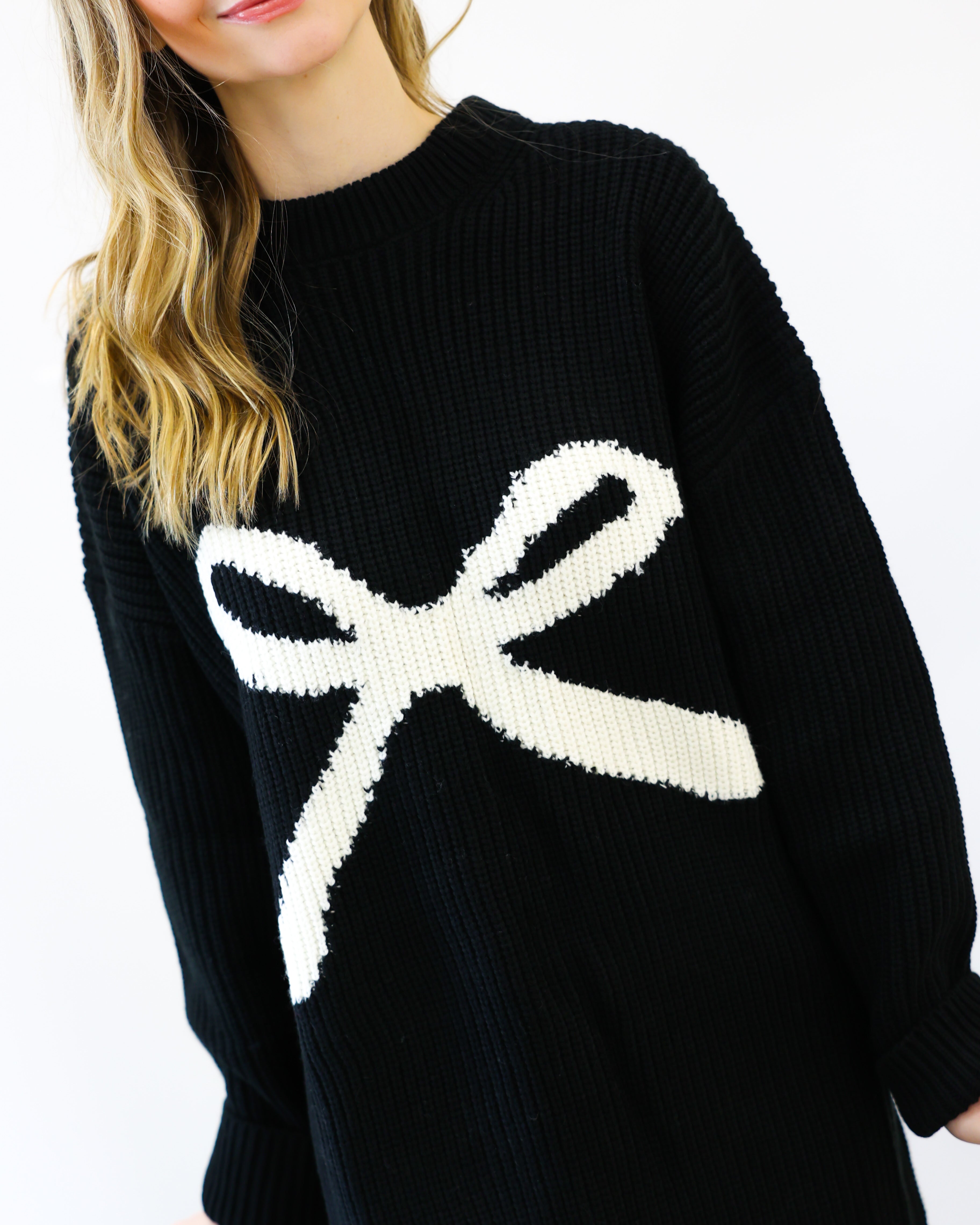 Black sweater with a large white bow design worn by a person on a white background