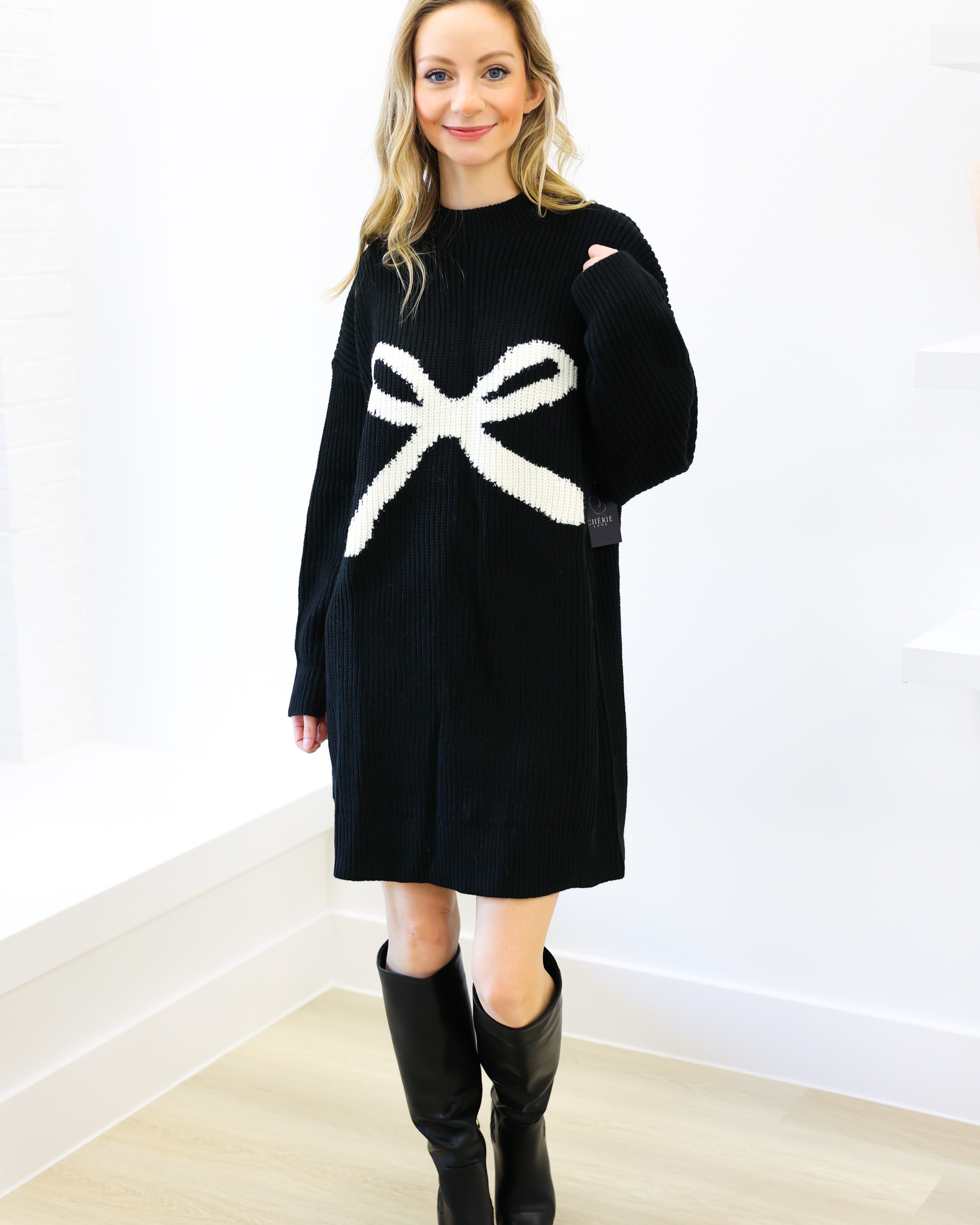 Woman wearing a black sweater dress with a white bow design, standing against a white wall.