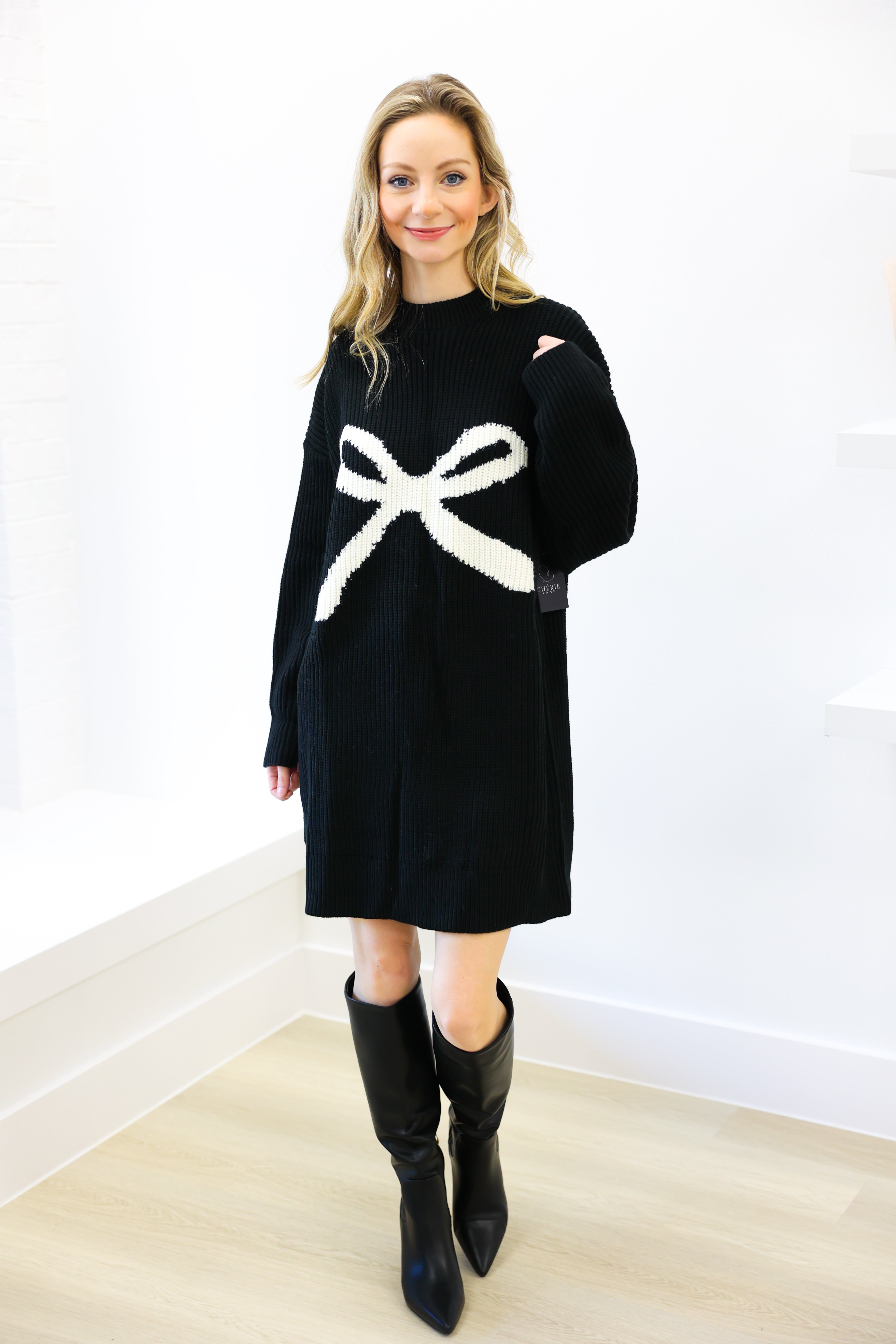 Woman wearing a black sweater dress with a white bow design, standing against a white wall.