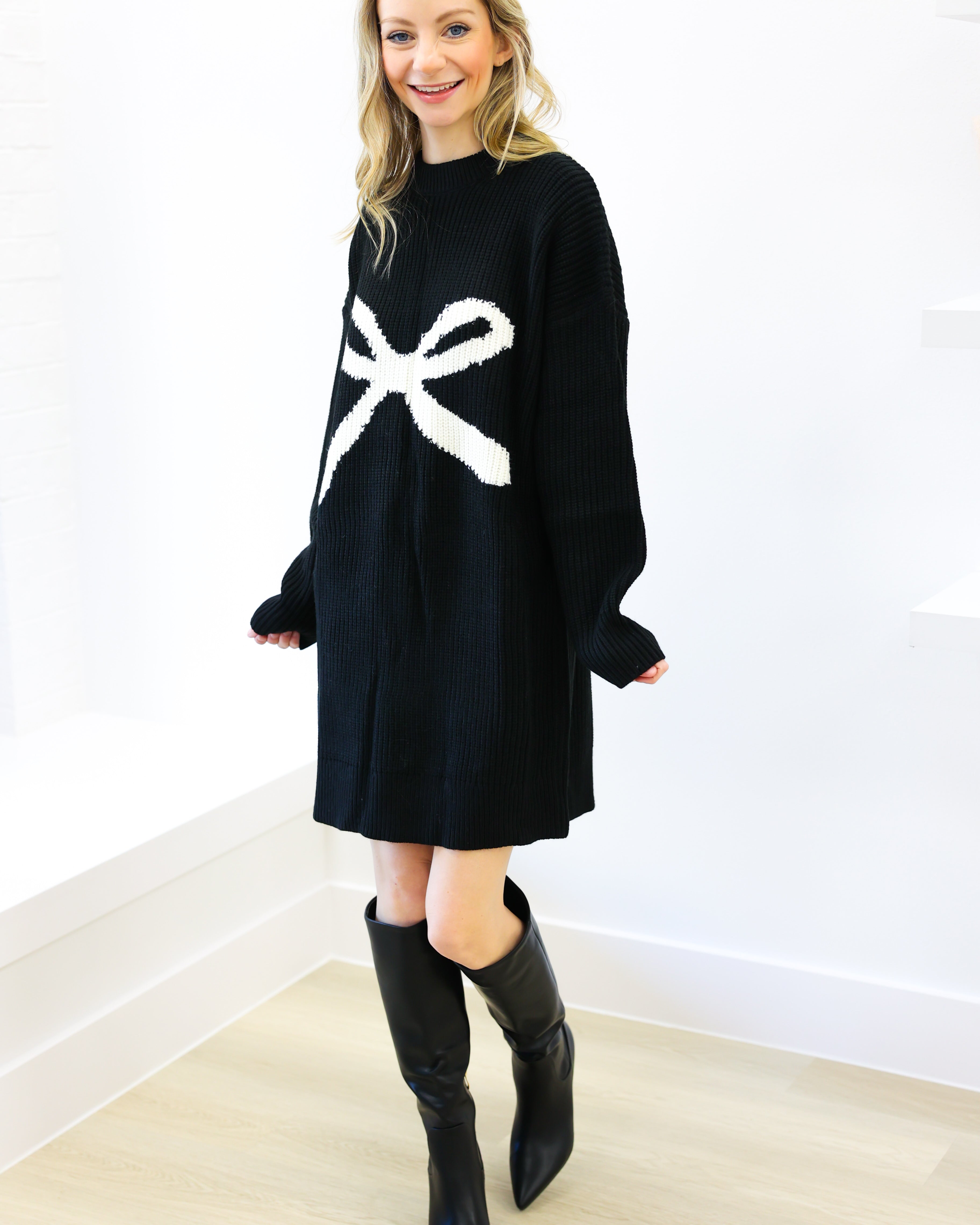 Woman wearing a black sweater dress with a white bow design and black knee-high boots on a white background.