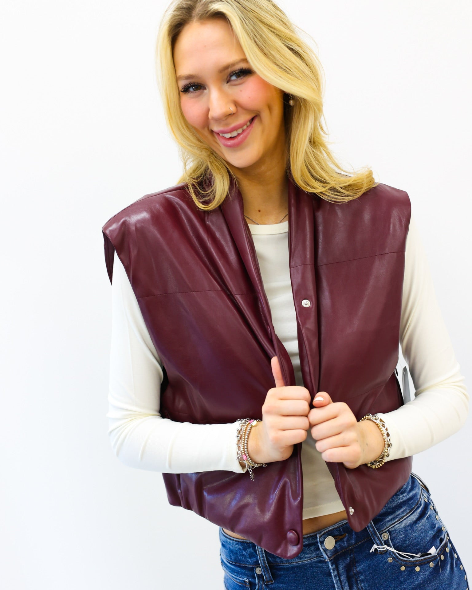 Woman wearing a maroon leather vest over a white long-sleeve shirt and blue jeans on a white background