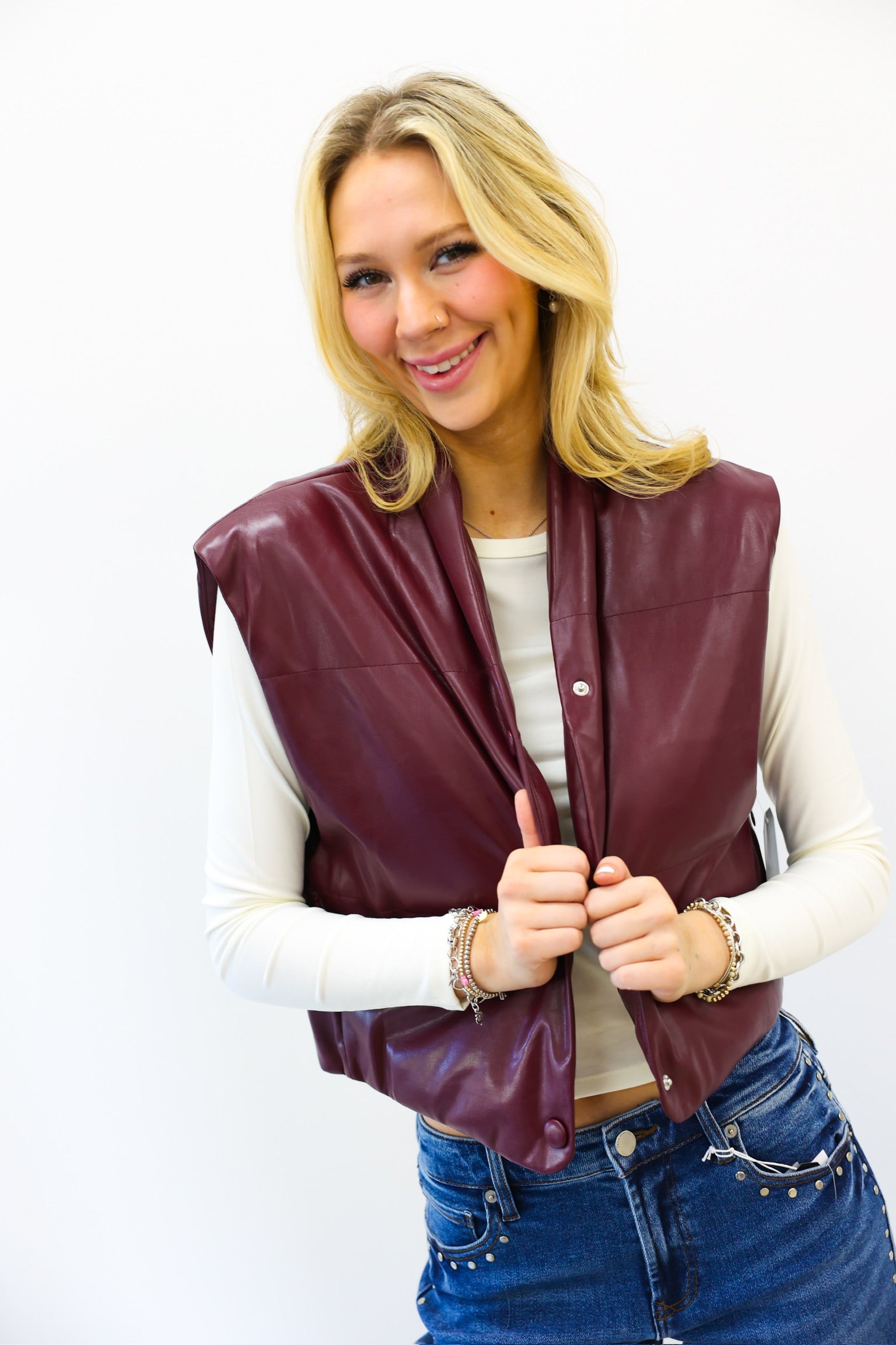 Woman wearing a maroon leather vest over a white long-sleeve shirt and blue jeans on a white background