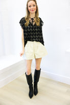 Woman wearing a black floral blouse, cream shorts, and black knee-high boots against a white wall.