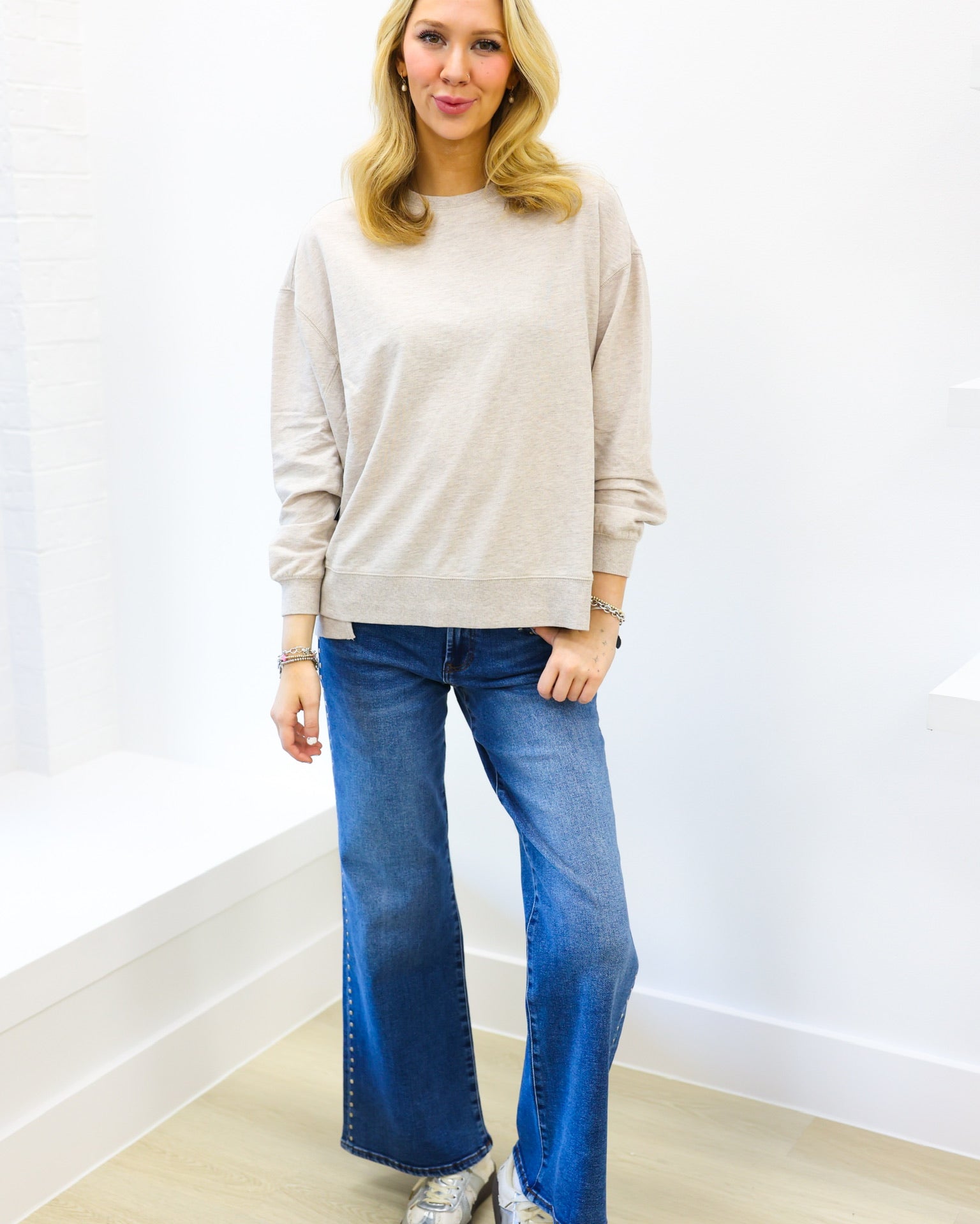 Woman wearing a beige sweater and blue jeans standing against a white wall.