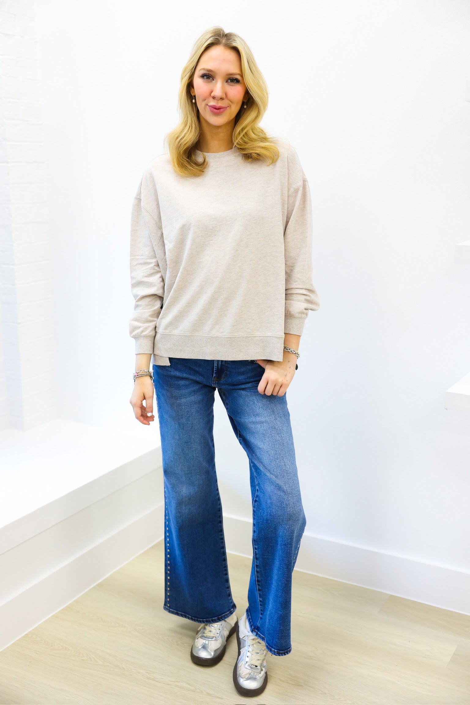 Woman wearing a beige sweater and blue jeans standing against a white wall.