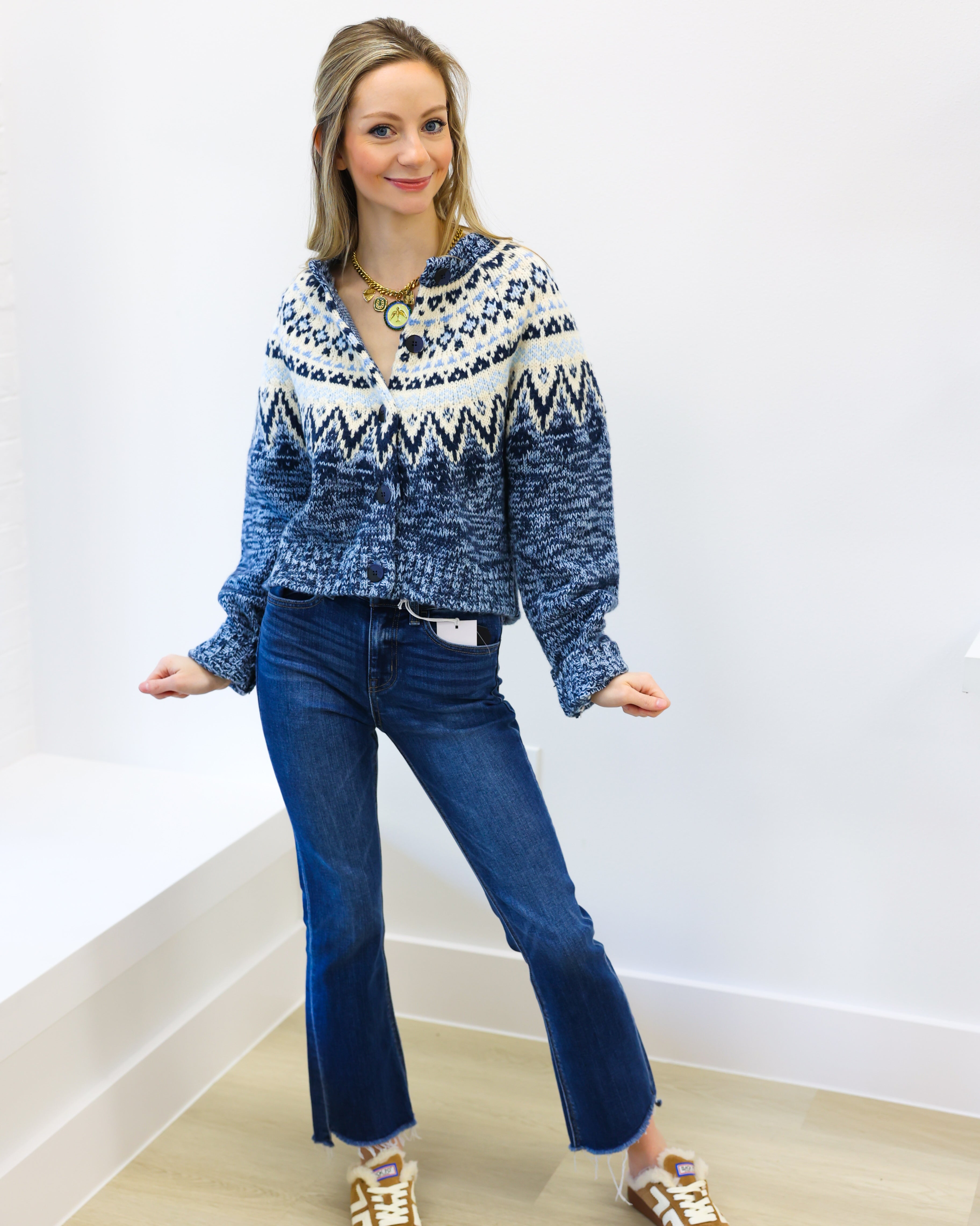 Woman wearing a blue patterned sweater and jeans standing against a white wall.