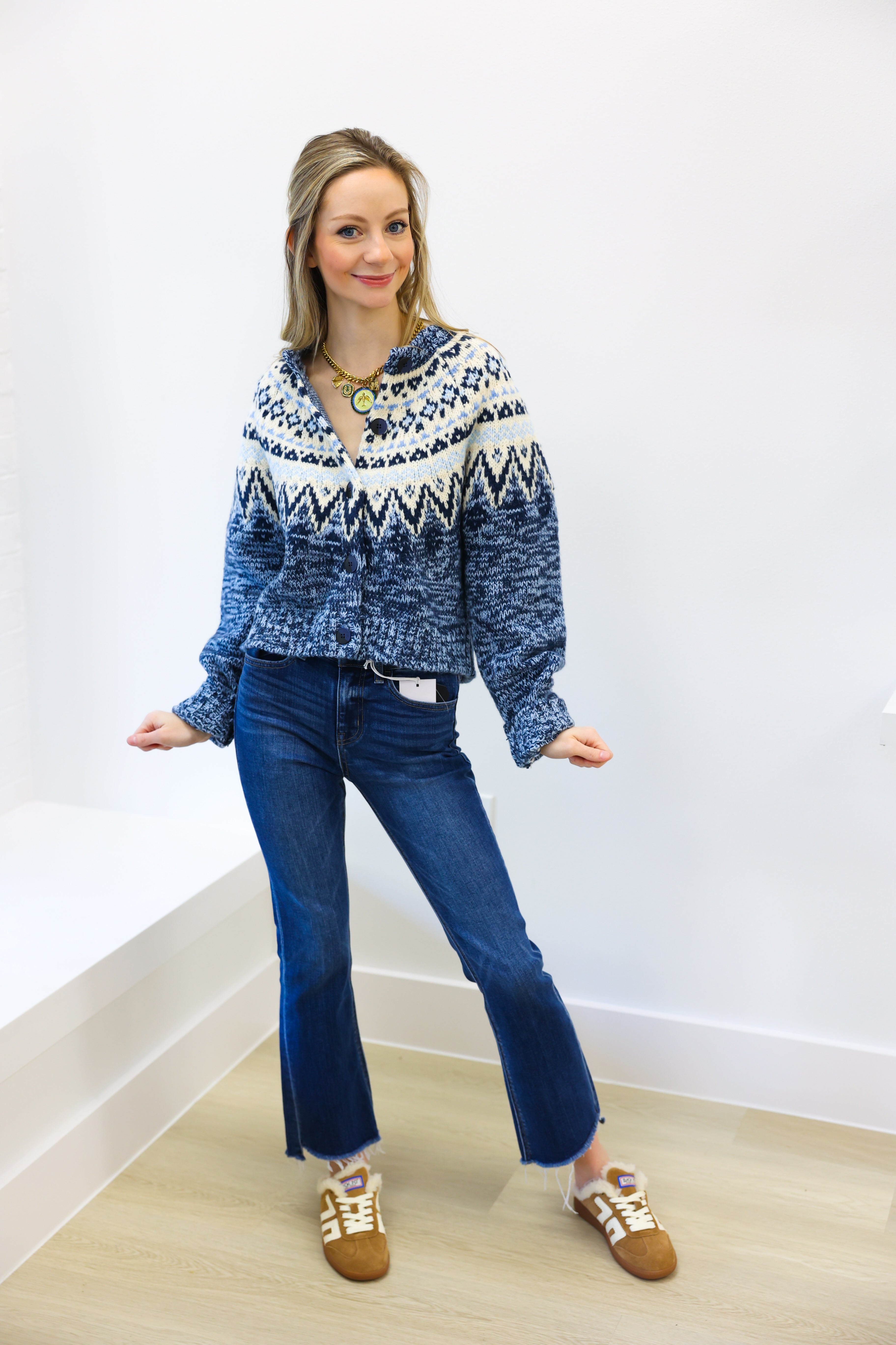 Woman wearing a blue patterned sweater and jeans standing against a white wall.