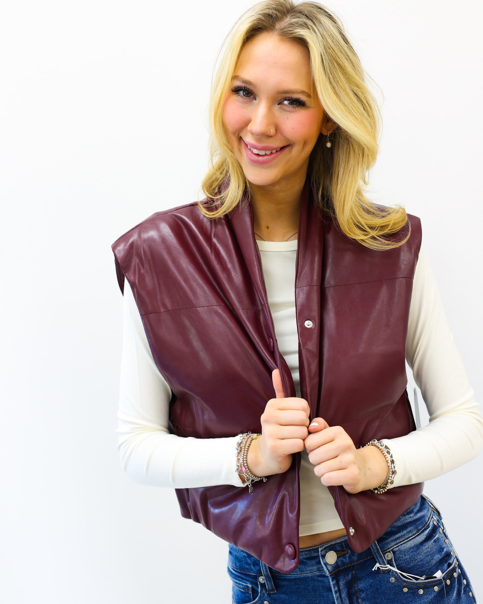 Woman wearing a burgundy leather vest over a white long-sleeve shirt and blue jeans on a white background
