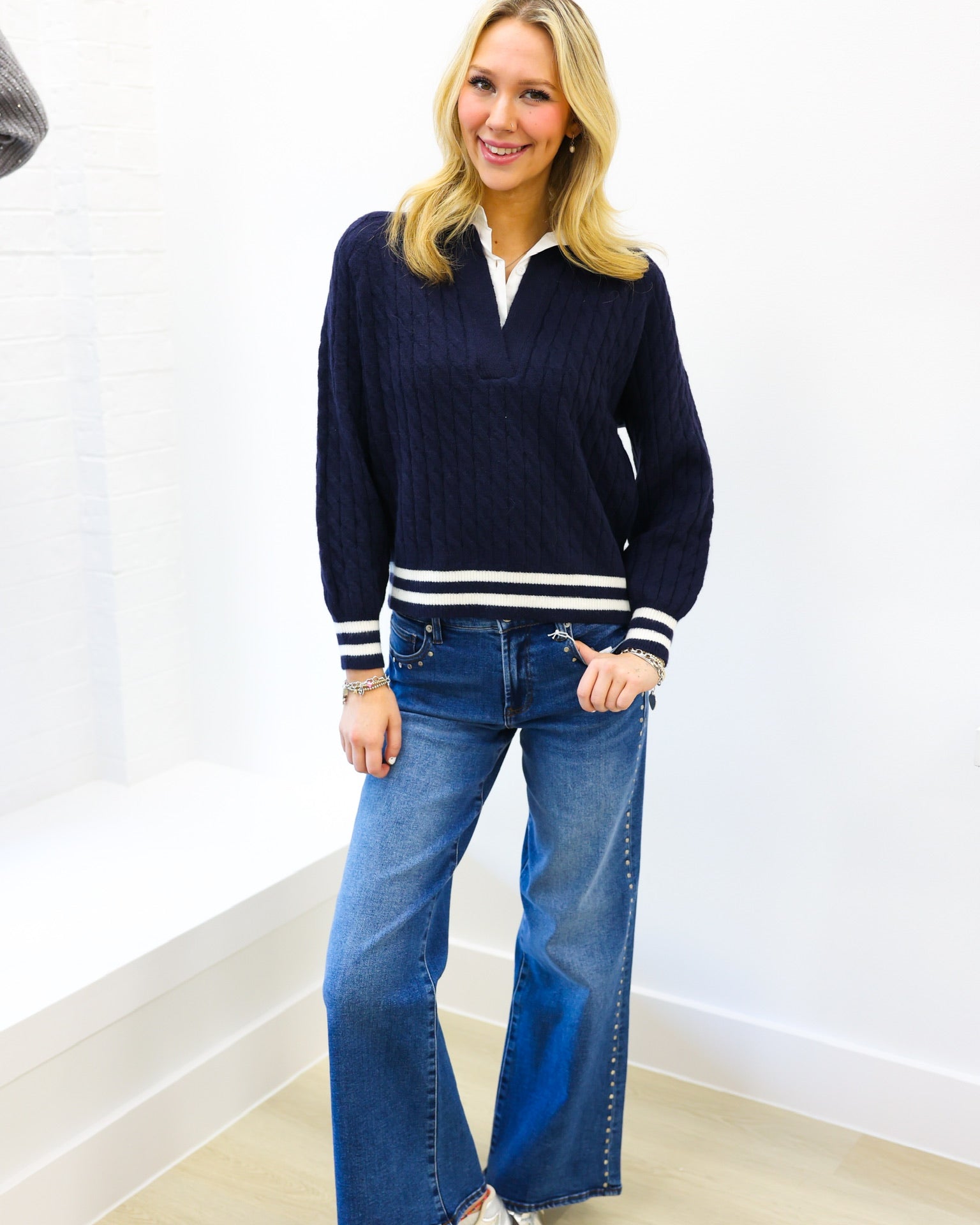 Woman wearing a navy sweater with white stripes and blue jeans against a white wall.