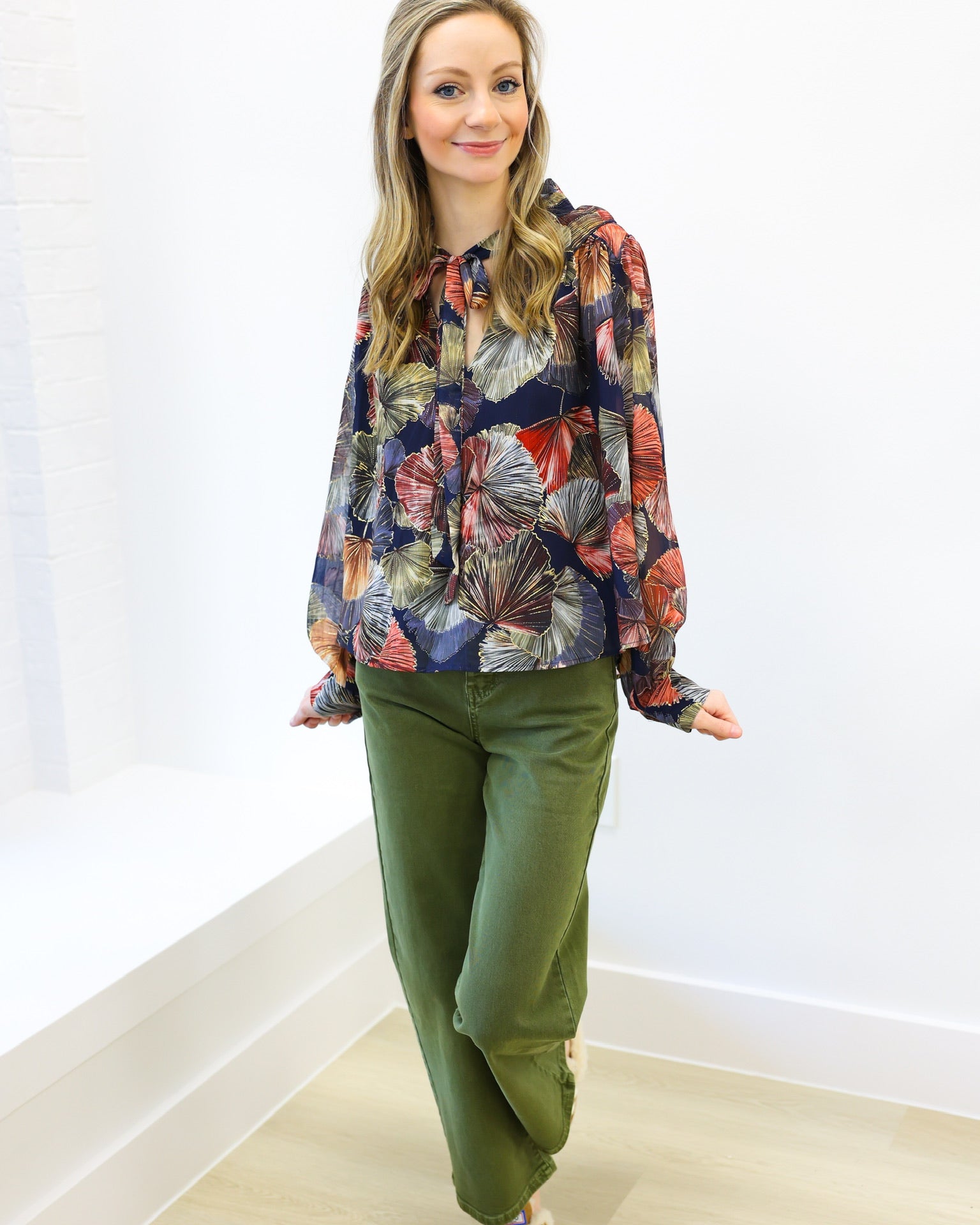 Woman wearing a colorful floral blouse and green pants against a white wall.