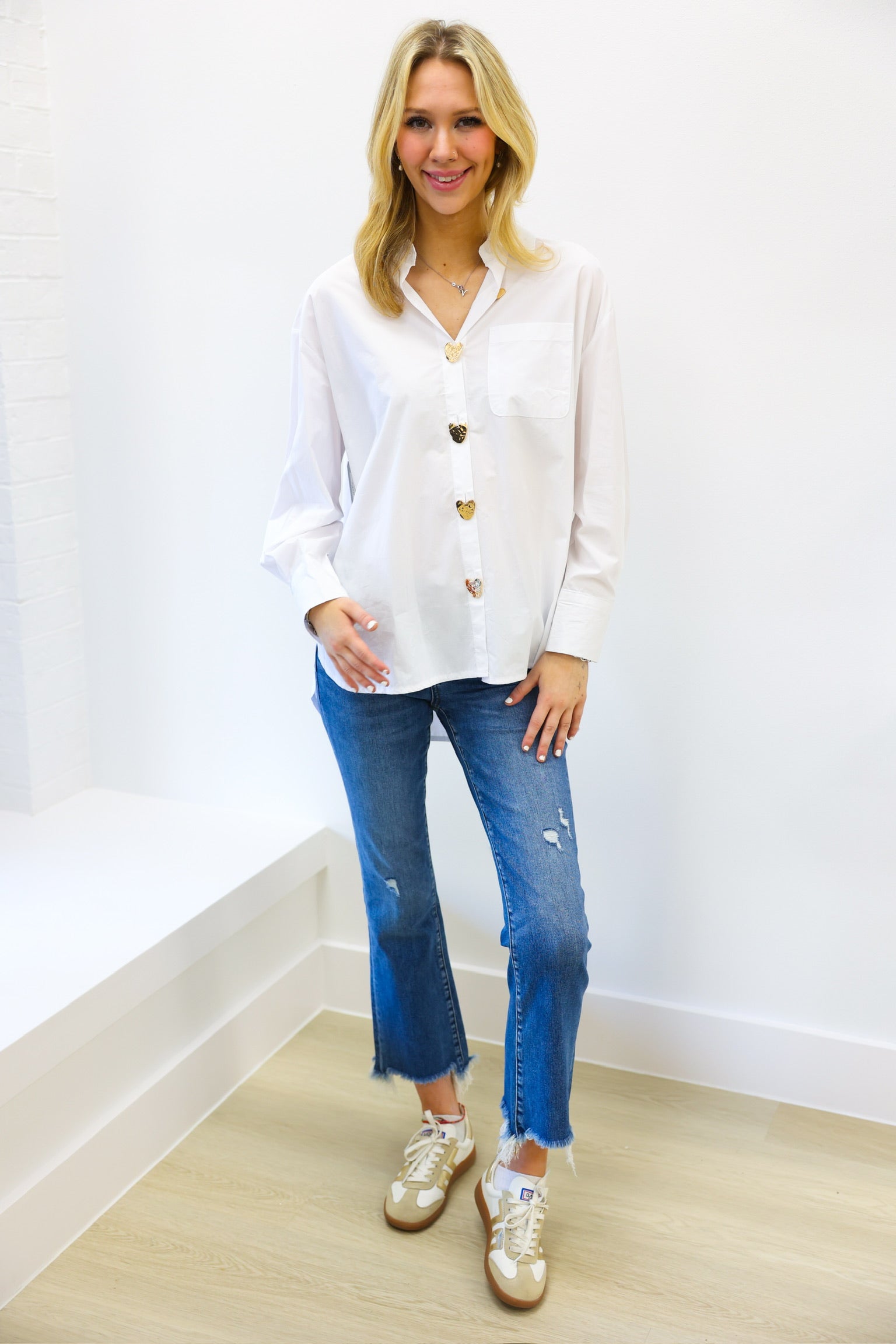 Woman wearing a white blouse with gold buttons, blue jeans, and white sneakers on a plain background