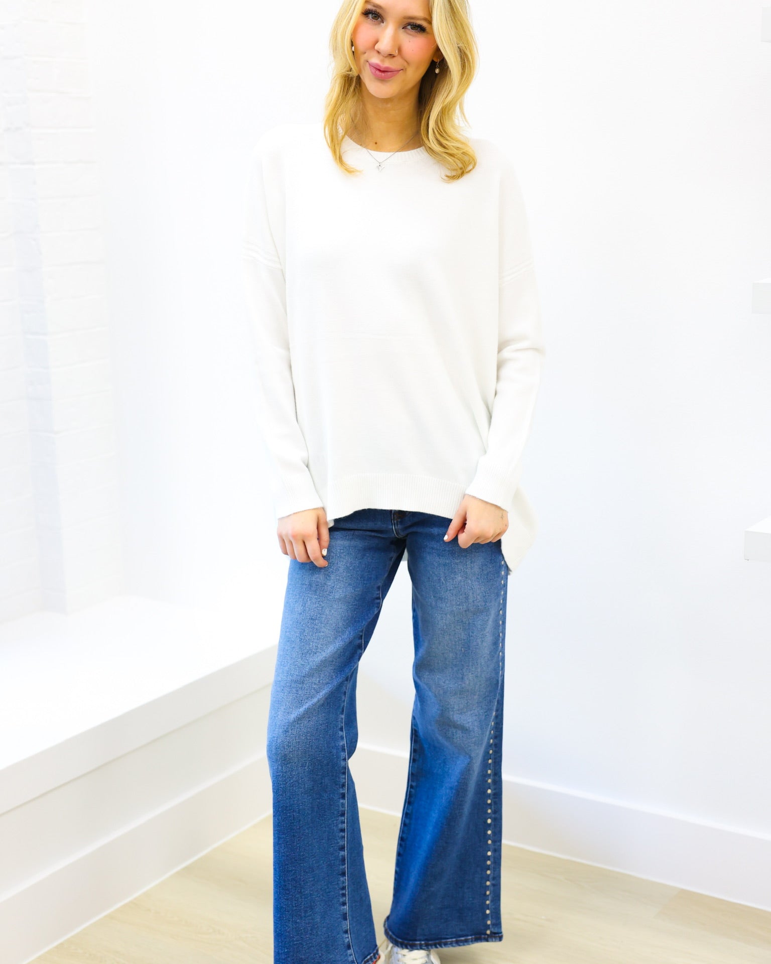 Woman wearing a white sweater and blue jeans standing against a white wall.