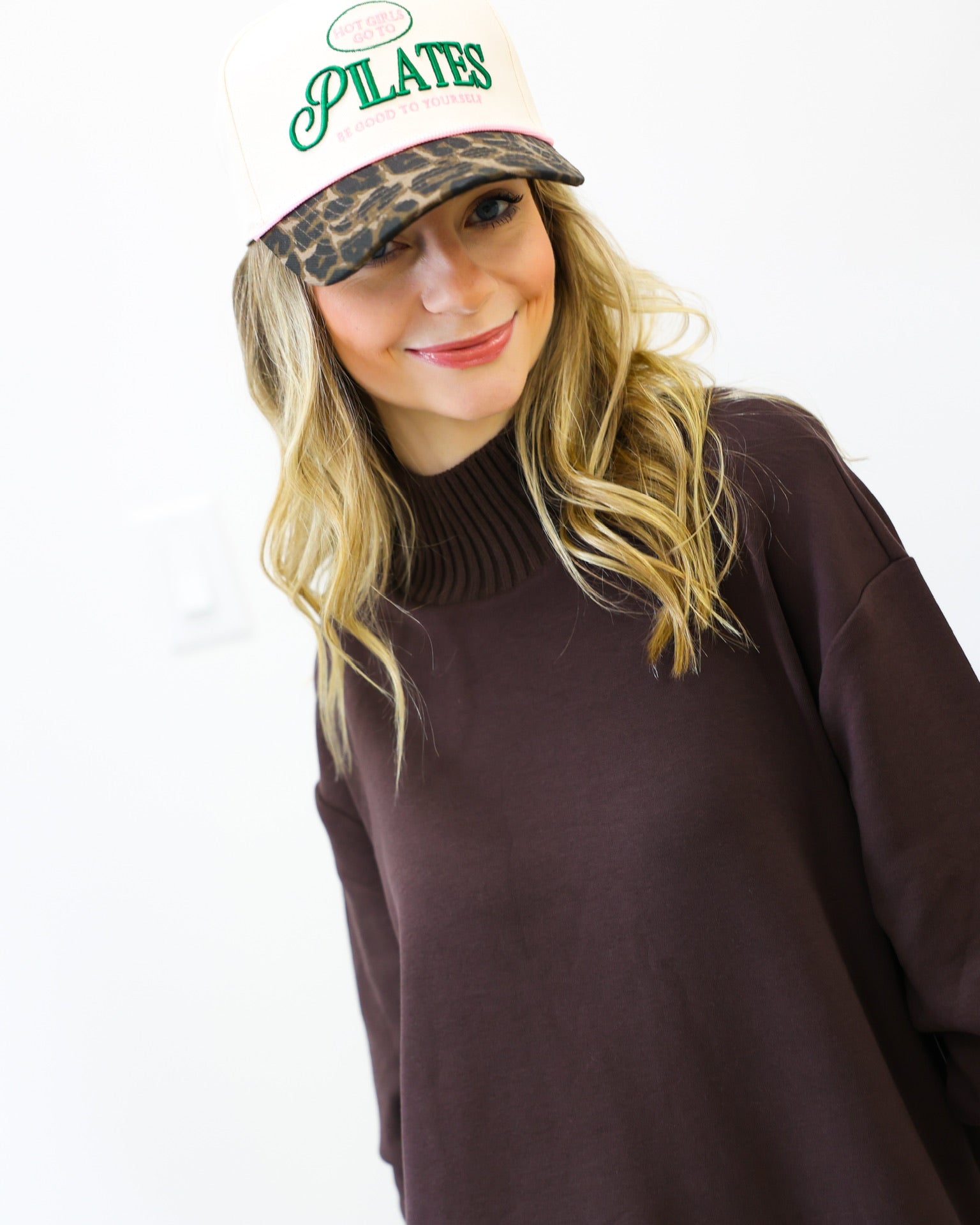 Woman wearing a brown sweater and a cap with 'Pilates' text on a white background