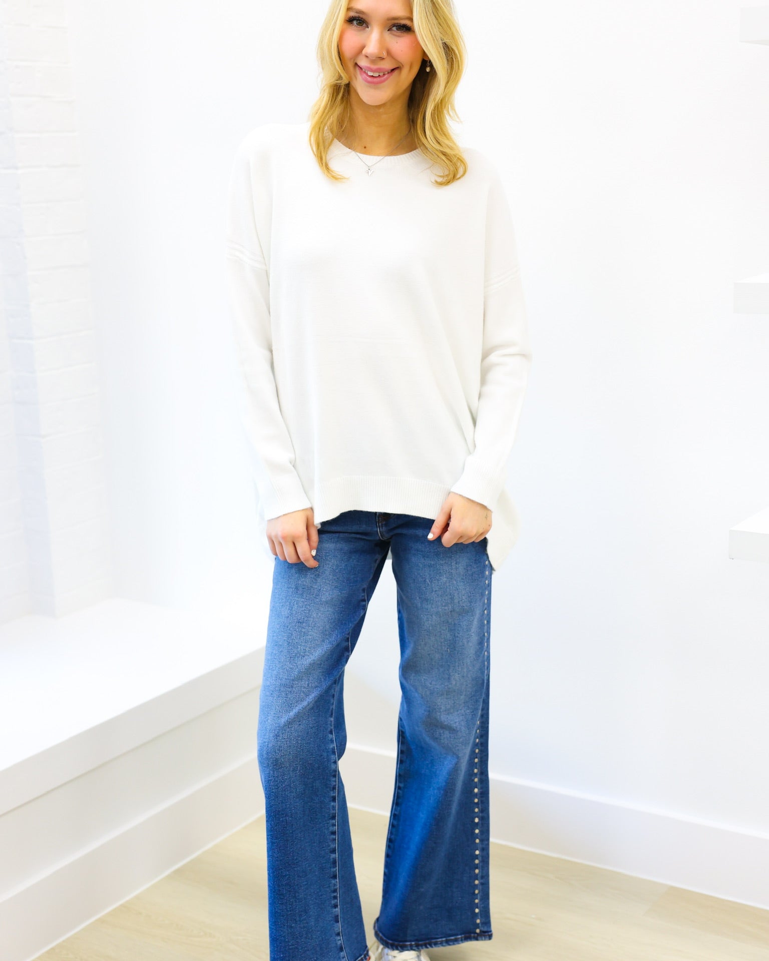 Woman wearing a white sweater and blue jeans standing against a white wall.