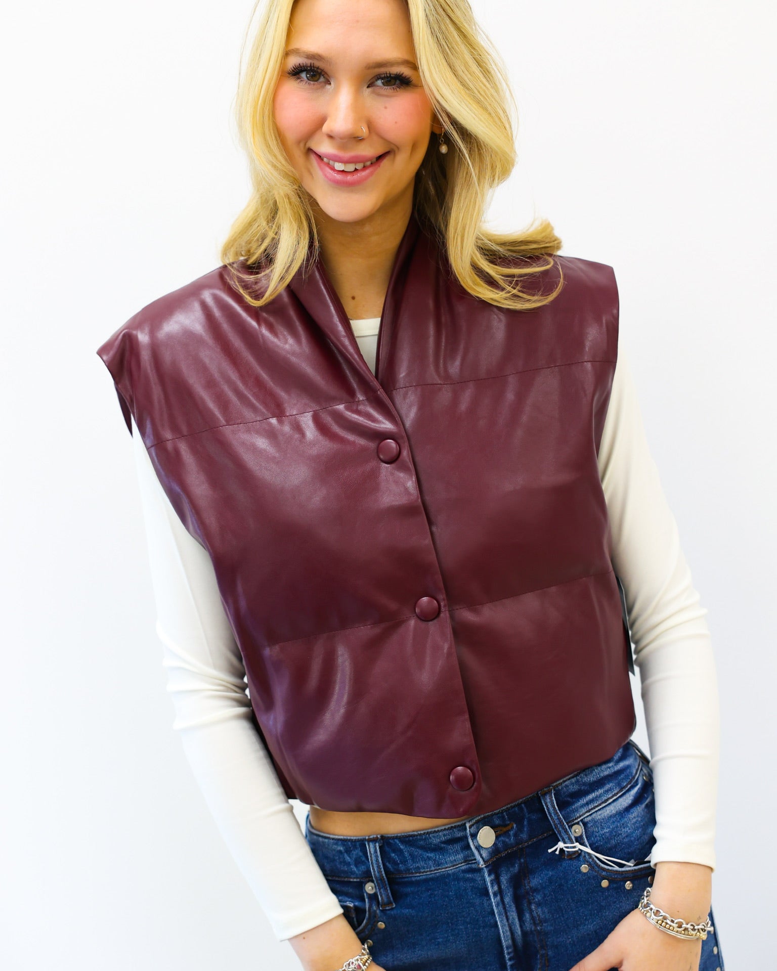 Woman wearing a maroon vest over a white long-sleeve shirt and blue jeans on a white background