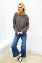 Woman wearing a gray sweater and blue jeans against a white wall.
