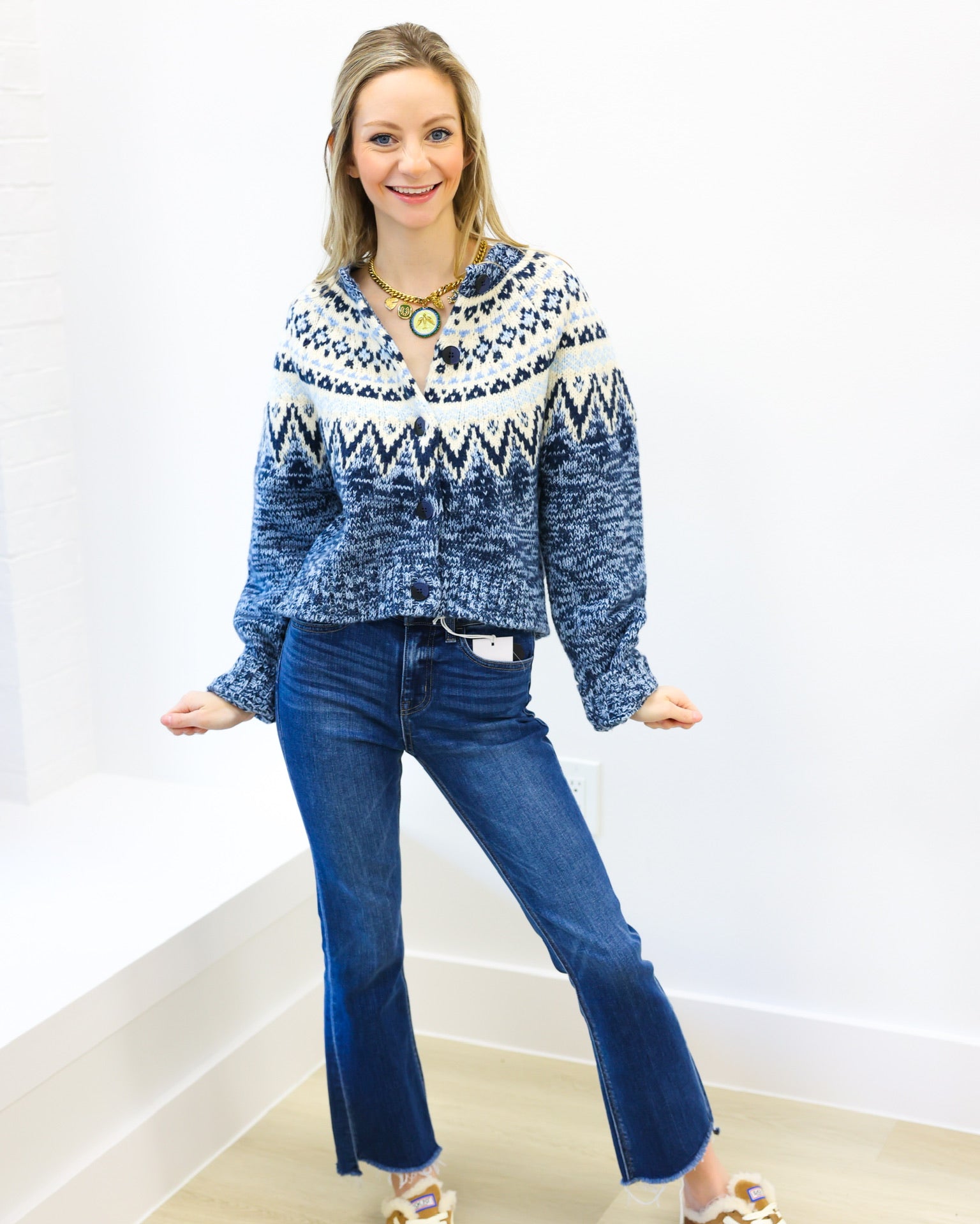 Woman wearing a blue patterned sweater and blue jeans standing against a white wall.