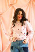 Woman wearing a pink sweater with red heart patterns against a peach-colored curtain.
