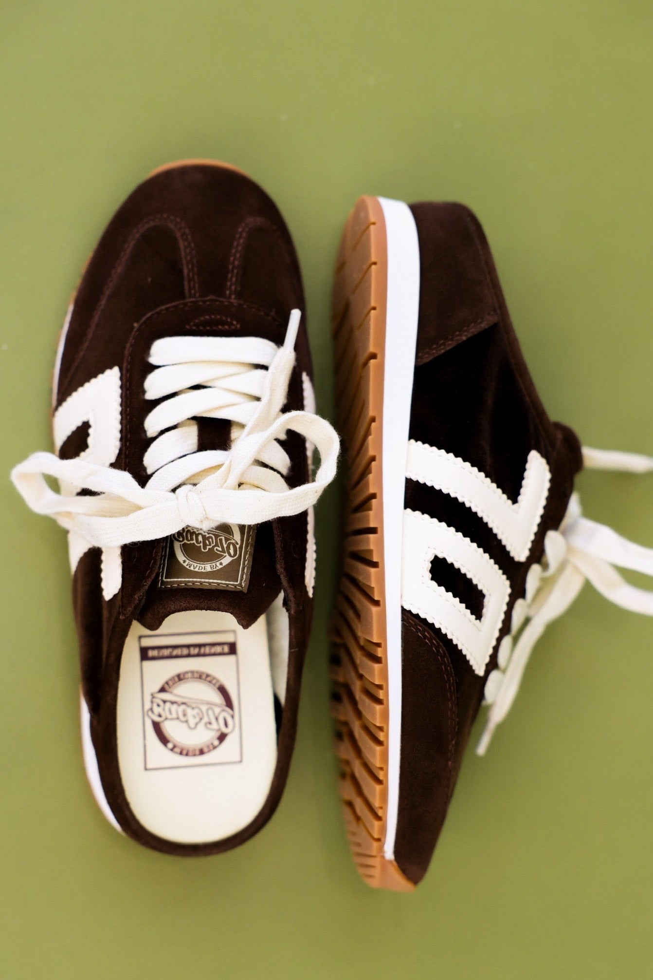 A pair of vintage-style low-top sneakers in dark brown suede with white stripes, white laces, and gum soles, displayed on a solid olive green background.
