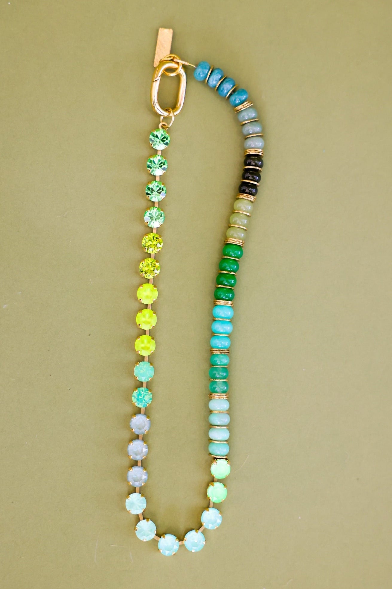 A beaded bracelet with a gradient of colorful round beads transitioning from pastel blue and purple to green, yellow, and turquoise, finished with a gold clasp on a solid beige background.