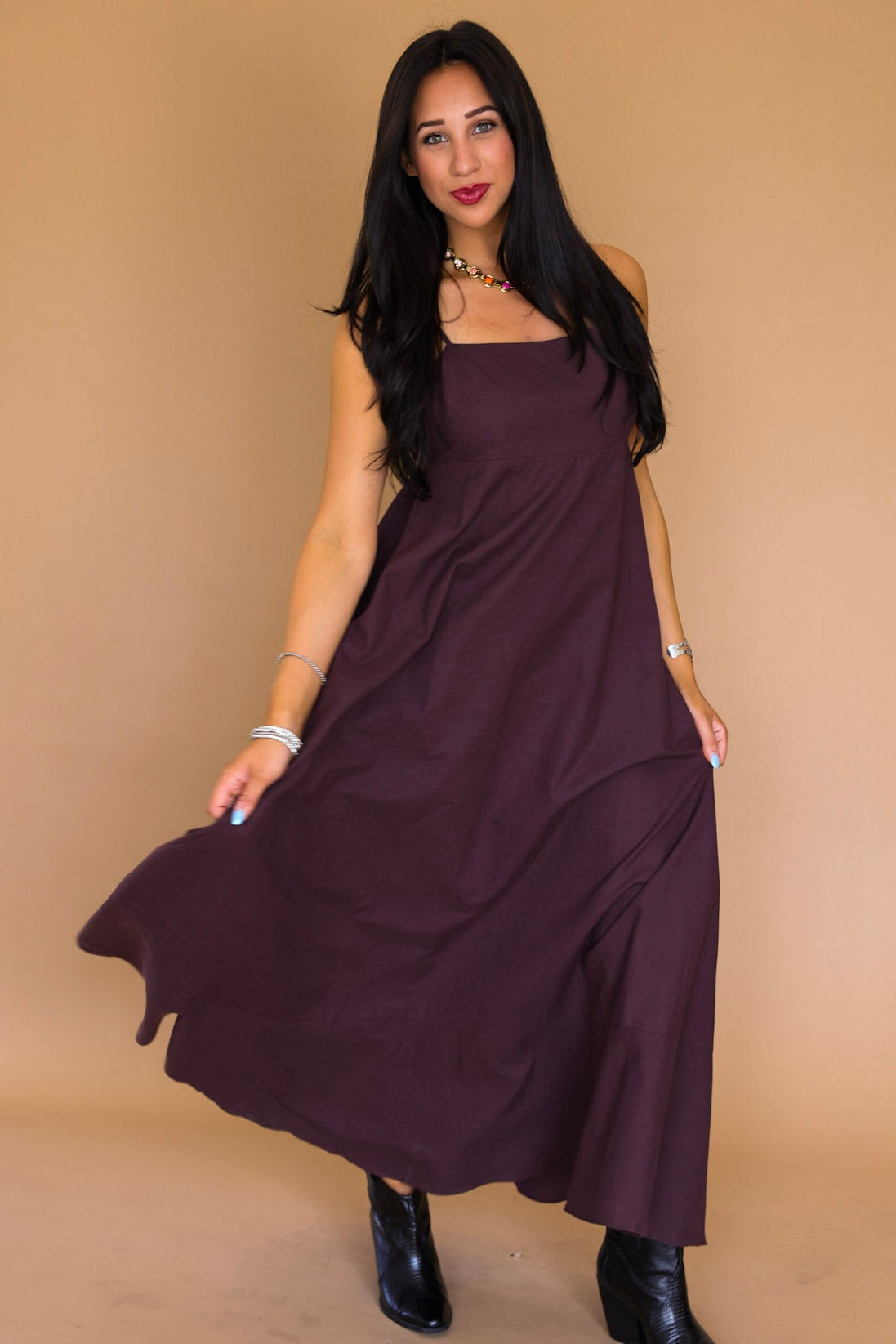 A woman modeling a sleeveless, flowy maxi dress in deep plum purple, paired with black ankle boots, standing against a solid tan studio backdrop.