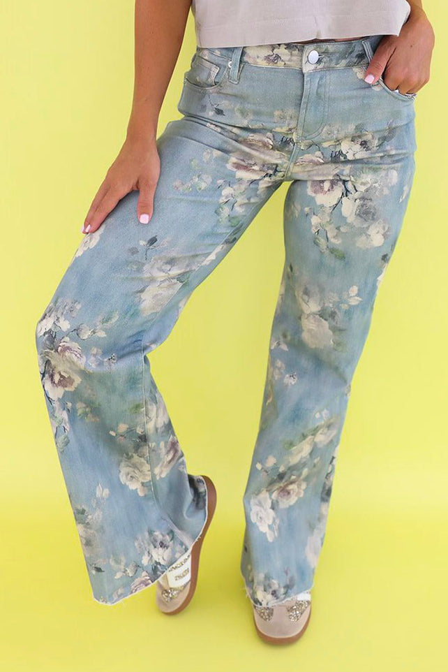 A woman modeling light blue floral wide-leg jeans with a white sleeveless top, standing against a solid pale yellow background.