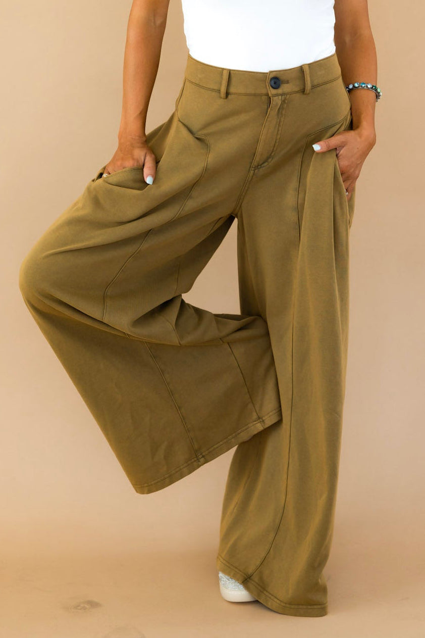 A woman posing in a white short-sleeve top and olive green wide-leg pants against a solid beige studio background.