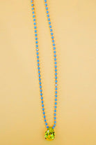 A beaded necklace featuring small light blue faceted beads and a large, faceted lime green teardrop pendant, displayed against a solid pale peach background.