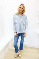 Woman wearing a light blue denim shirt and jeans standing against a white wall.