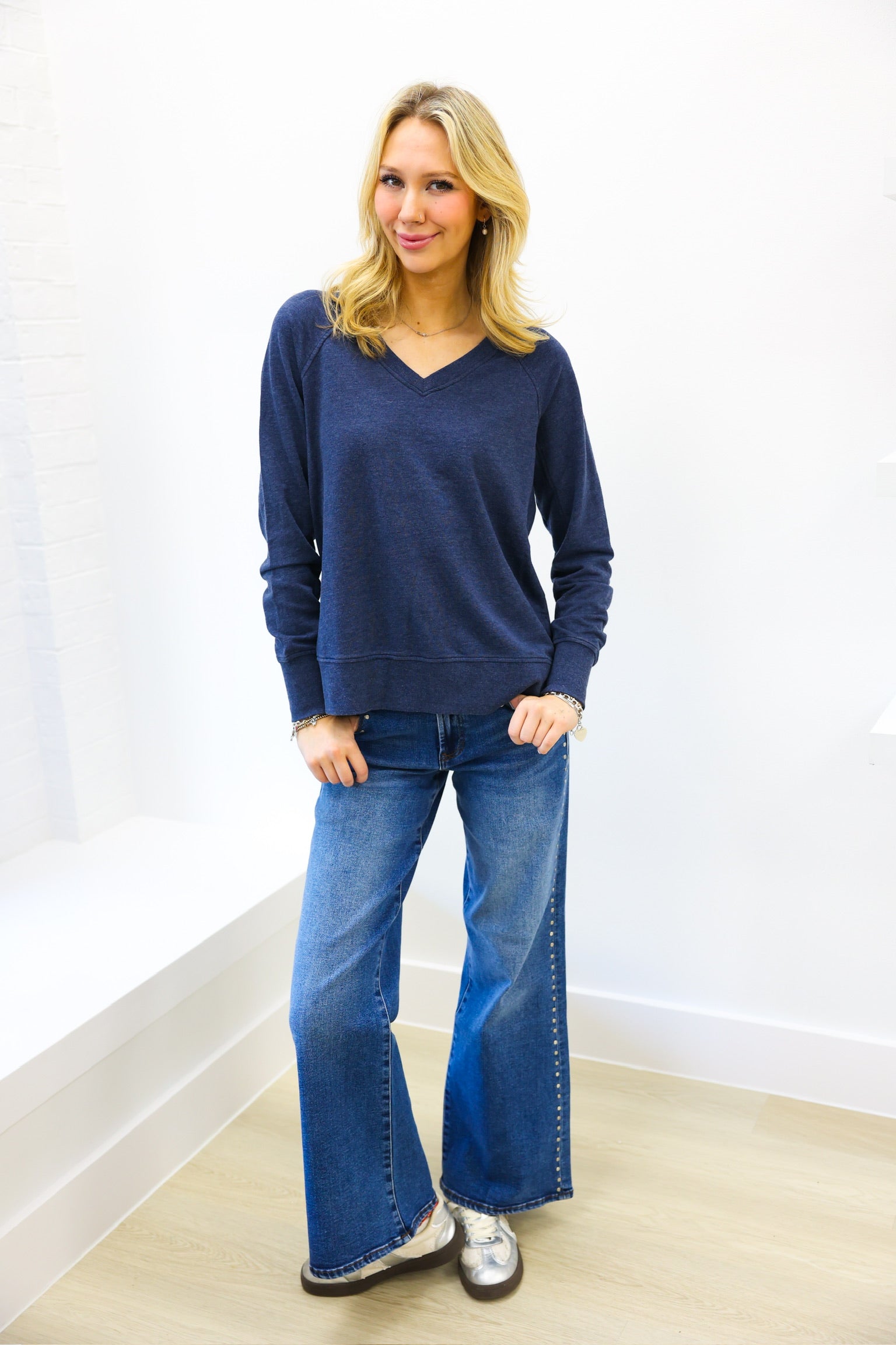 Woman wearing a blue sweater and jeans standing against a white wall.