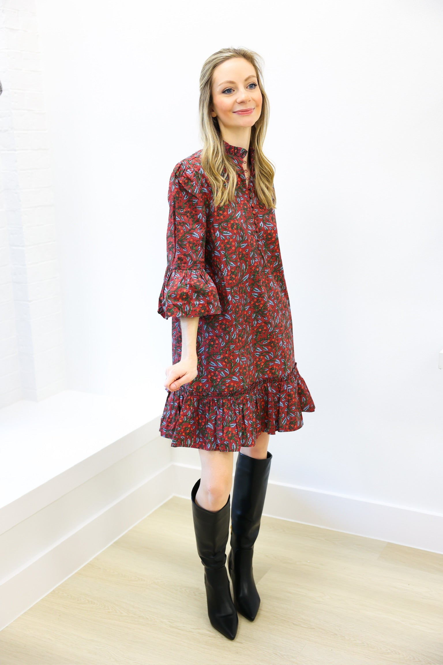 Woman wearing a red floral dress and black boots against a white wall.