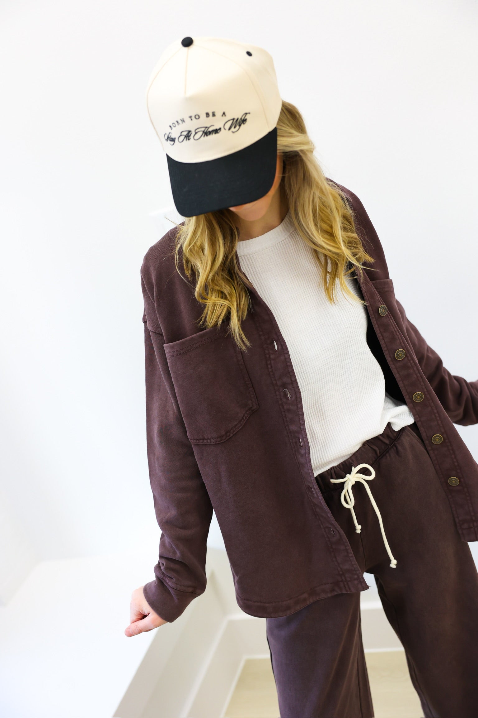 Person wearing a brown jacket, white shirt, and purple pants with a cap on a white background