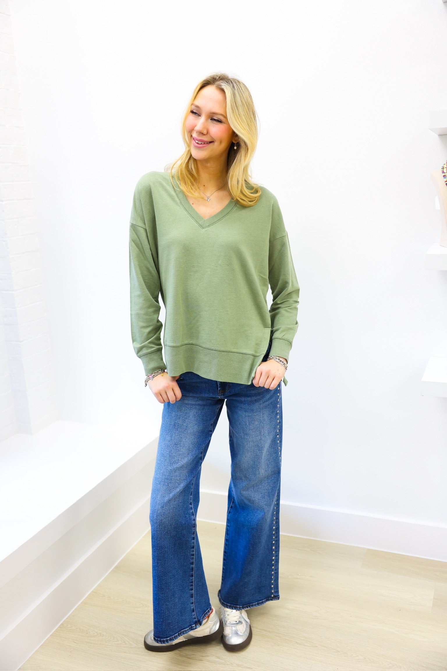 Woman wearing a green sweater and blue jeans standing against a white wall.