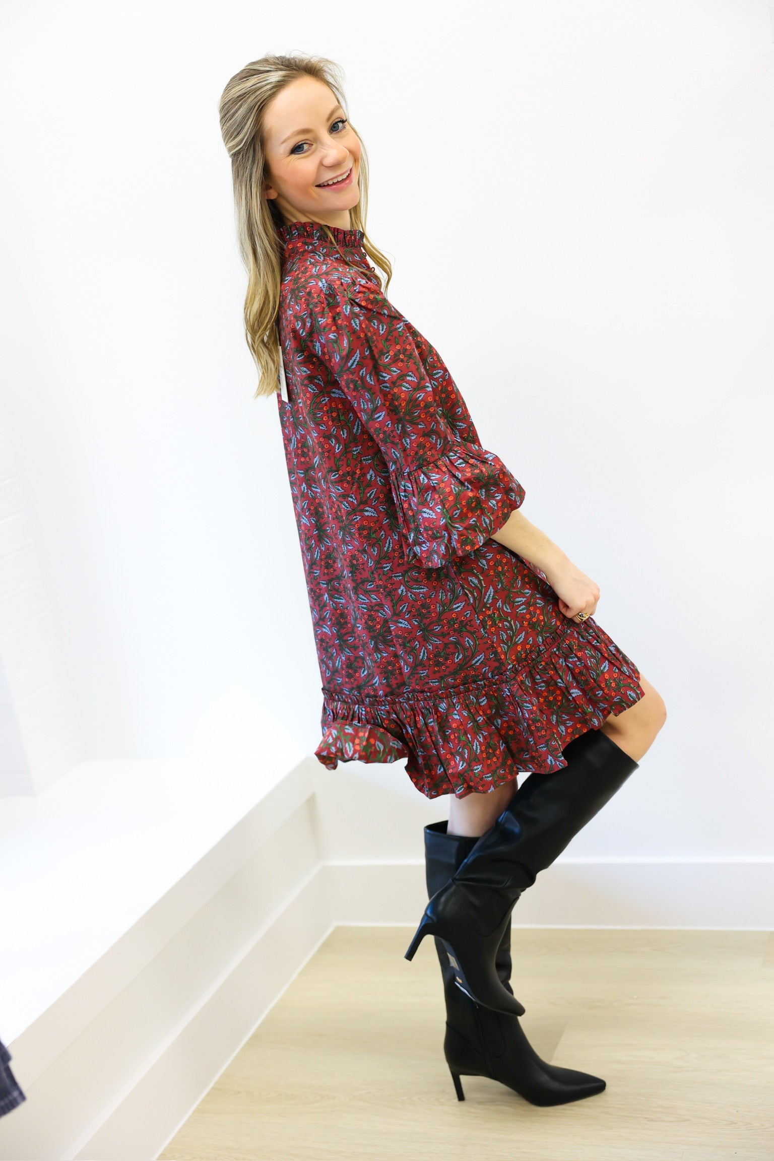 Woman wearing a red floral dress and black knee-high boots against a white wall.