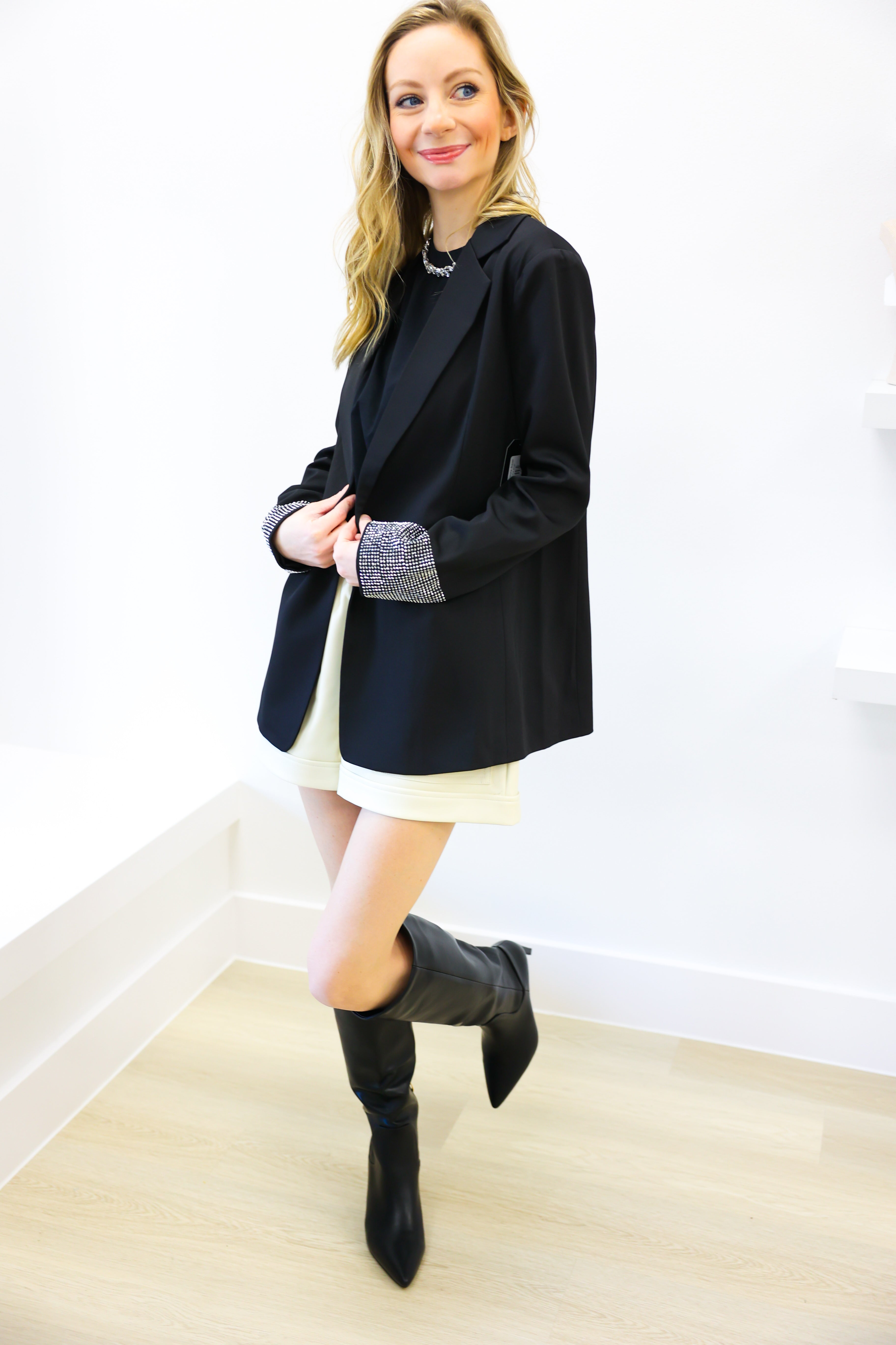 Woman wearing a black blazer, white dress, and black boots against a white wall.