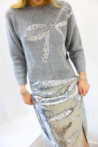 Person wearing a gray sweater with a large sequin bow and a silver sequin skirt on a white background